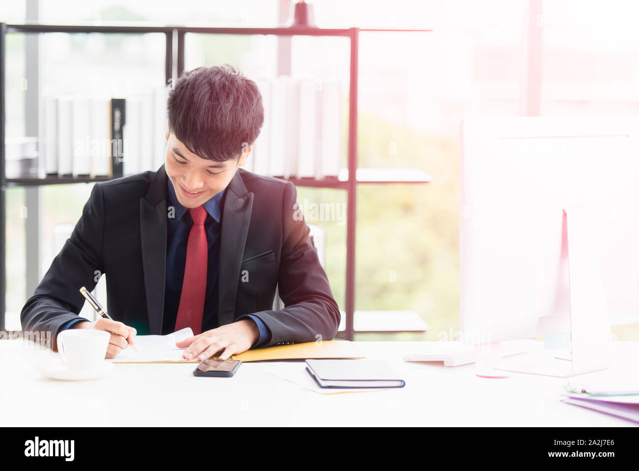 Un jeune homme asiatique porte un costume est assis la signature de documents papier avec un stylo sur un bureau dans le bureau. Il sourit et heureusement parce que le grand pr Banque D'Images