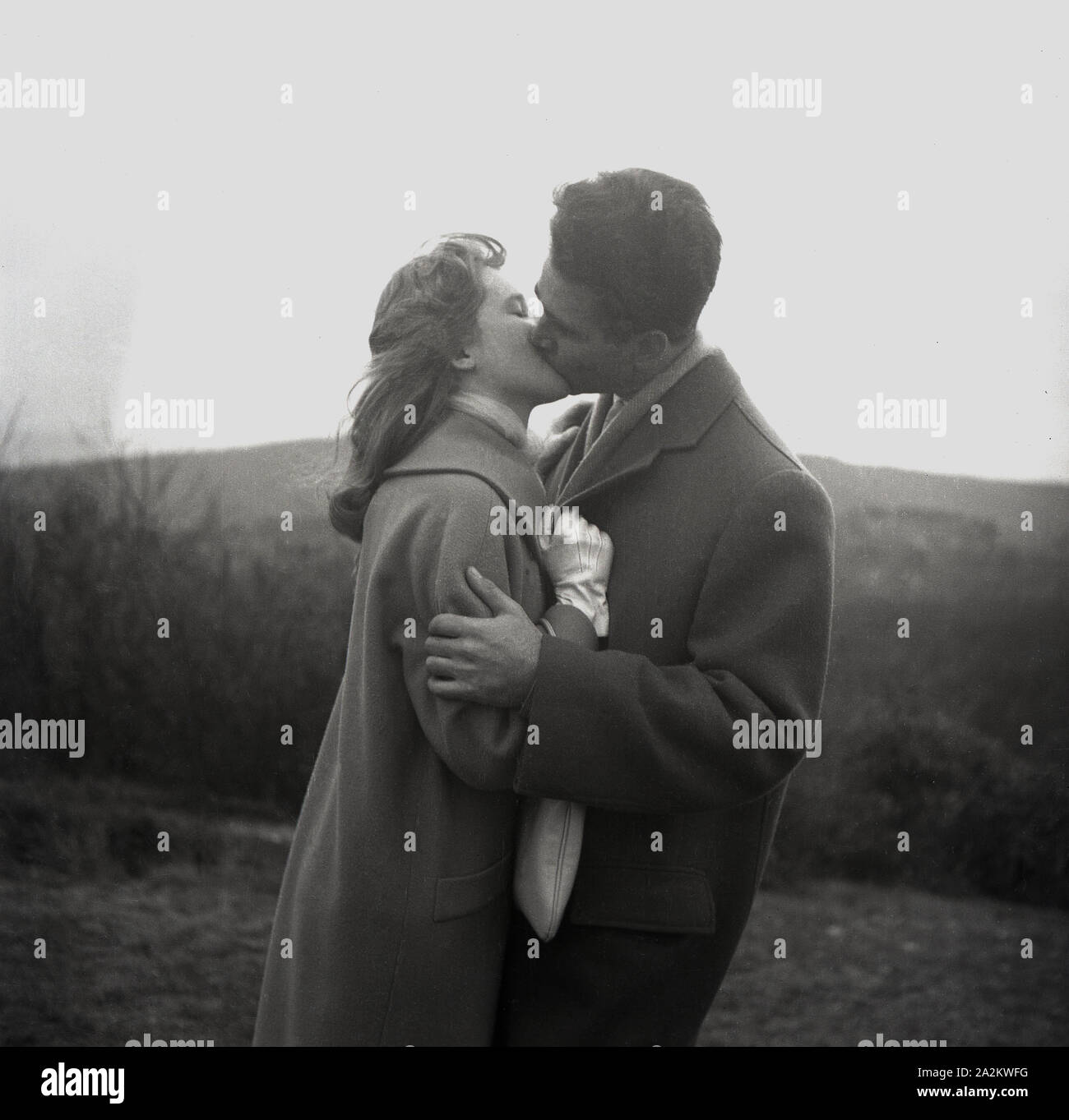 Années 1960, historique, deux amoureux s'embrassant, un homme et une femme portant des manteaux à l'extérieur sur une colline ayant une romance, Angleterre, Royaume-Uni. Banque D'Images