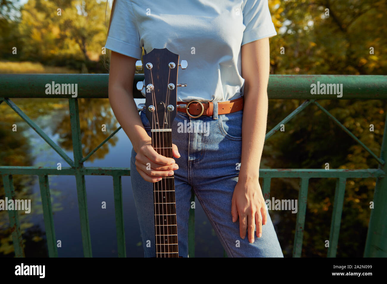 Jeune fille se tient sur le pont près de la clôture. Femme tenant une guitare acoustique par le manche de la guitare. La belle nature et rivière sur l'arrière-plan. Banque D'Images