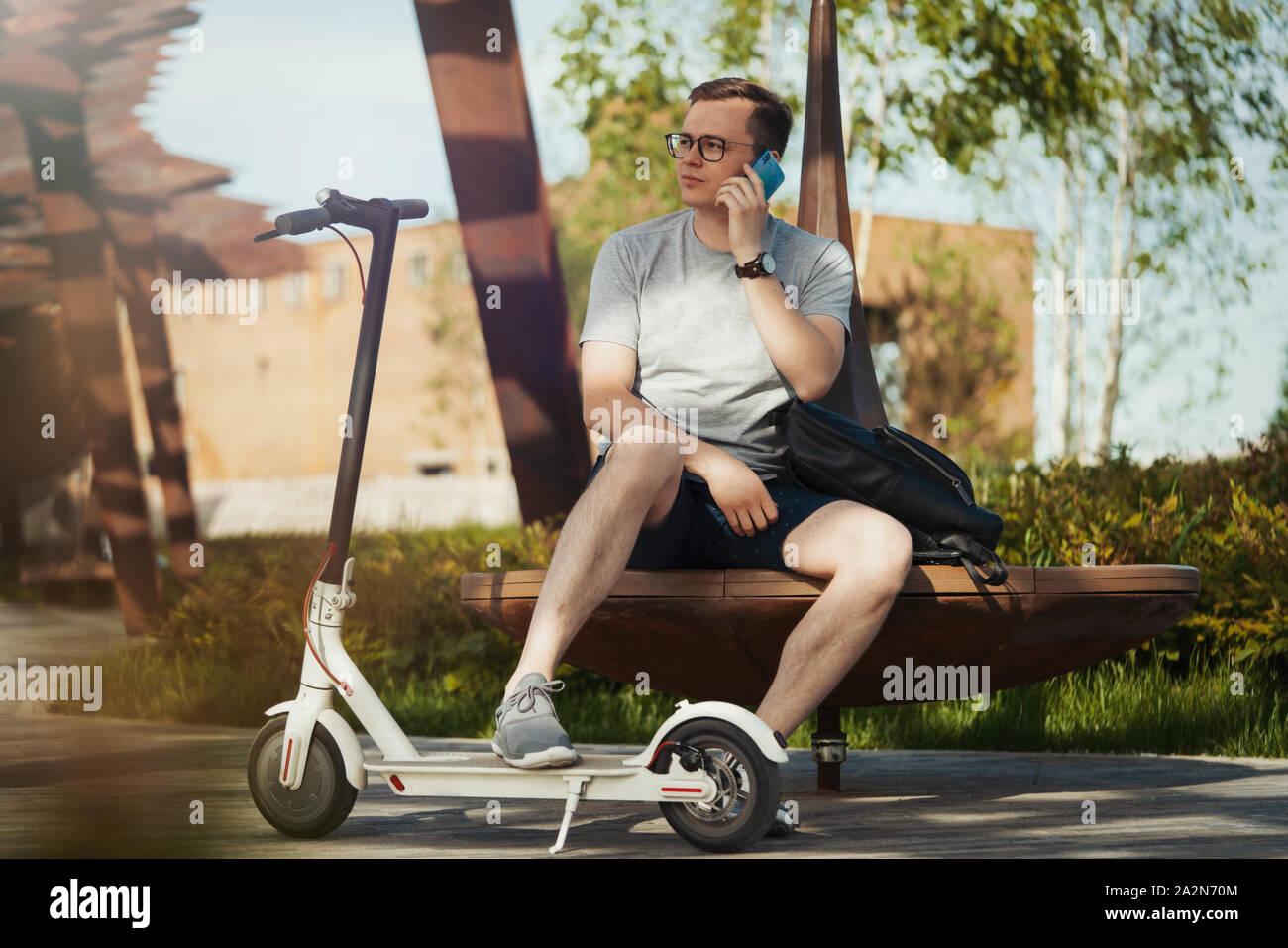 Homme séduisant de parler par téléphone mobile et assis à côté d'electric scooter de coup au paysage du parc moderne. L'homme est sur le premier plan, parc moderne est sur bac Banque D'Images
