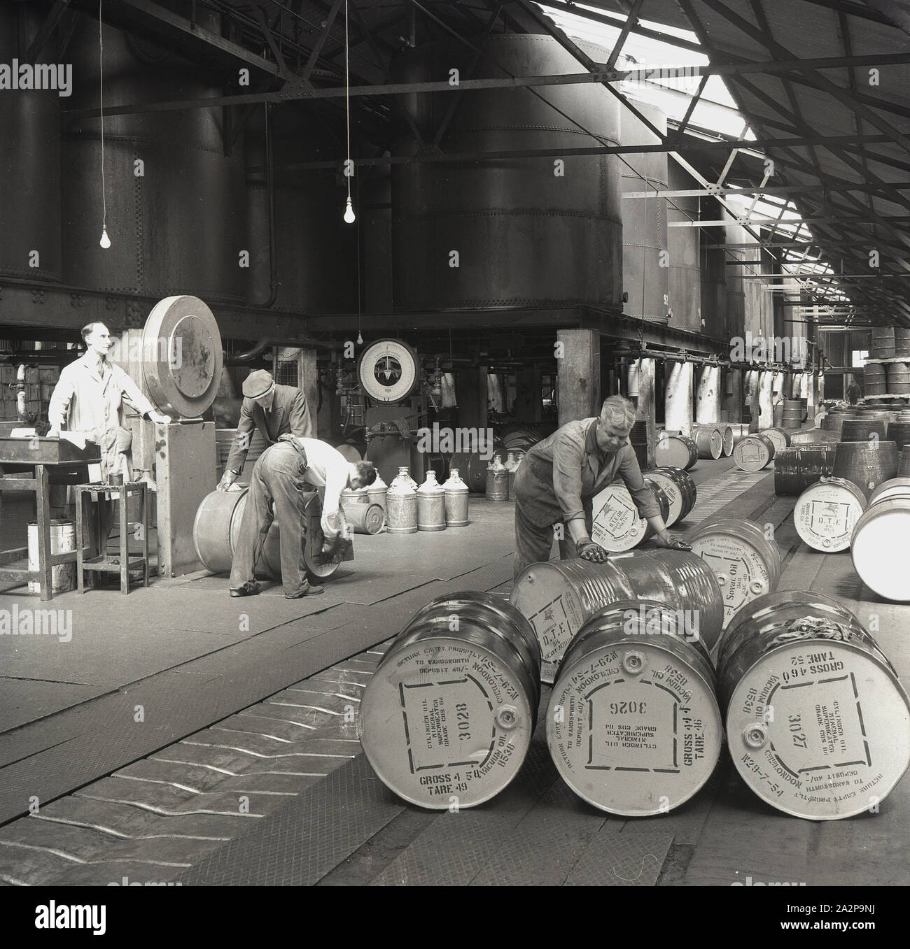 Années 1950, historiques, travailleurs masculins à l'intérieur d'un entrepôt remplissant des fûts d'huile moteur, Angleterre, Royaume-Uni. Des cycleurs géants reposent sur des poteaux en béton dans une partie du bâtiment. Banque D'Images