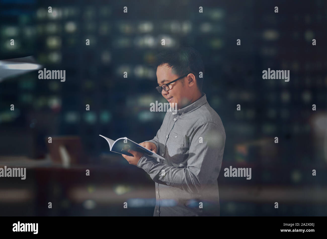 Jeune homme à la recherche heureux et souriant alors qu'il est en train de lire un livre. Student wearing à manches longues et des lunettes de donner les émotions positives tout en lisant un livre Banque D'Images