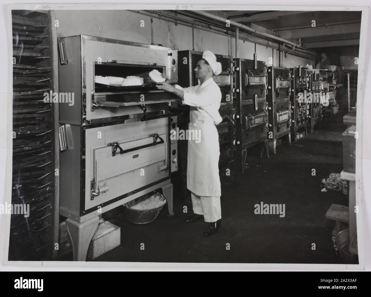 Photographie - Electrics Hecla Pty Ltd, fours à utiliser en cuisine industrielle, vers 1930, photographie en noir et blanc d'une cuisine industrielle doté d''une longue rangée de fours électriques à Hecla vers 1930. Un chef est au premier plan. Cette photographie est d'un album contenant 255 photographies en noir et blanc illustrant les appareils électriques, d'exposition, affiche des intérieurs de l'usine et le matériel publicitaire portant sur l'île Hecla Electrics Pty Ltd. Il fait partie de la collection de photographies d'Hecla Banque D'Images