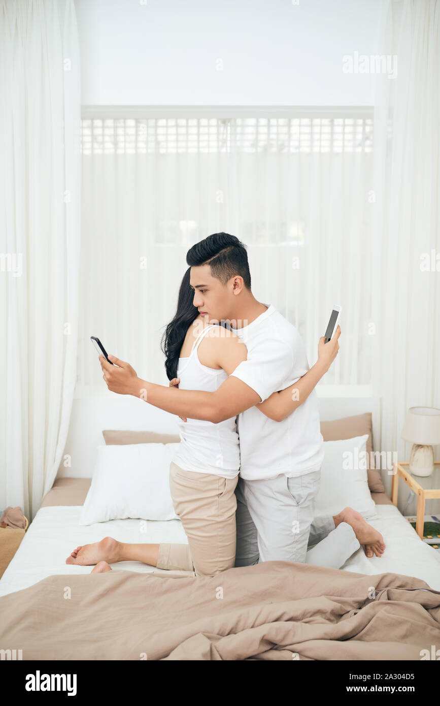 Jeune couple dans la chambre à coucher. Vue de côté de l'homme et de la femme infidèle sont tricher sur le téléphone tout en serrant l'autre Banque D'Images