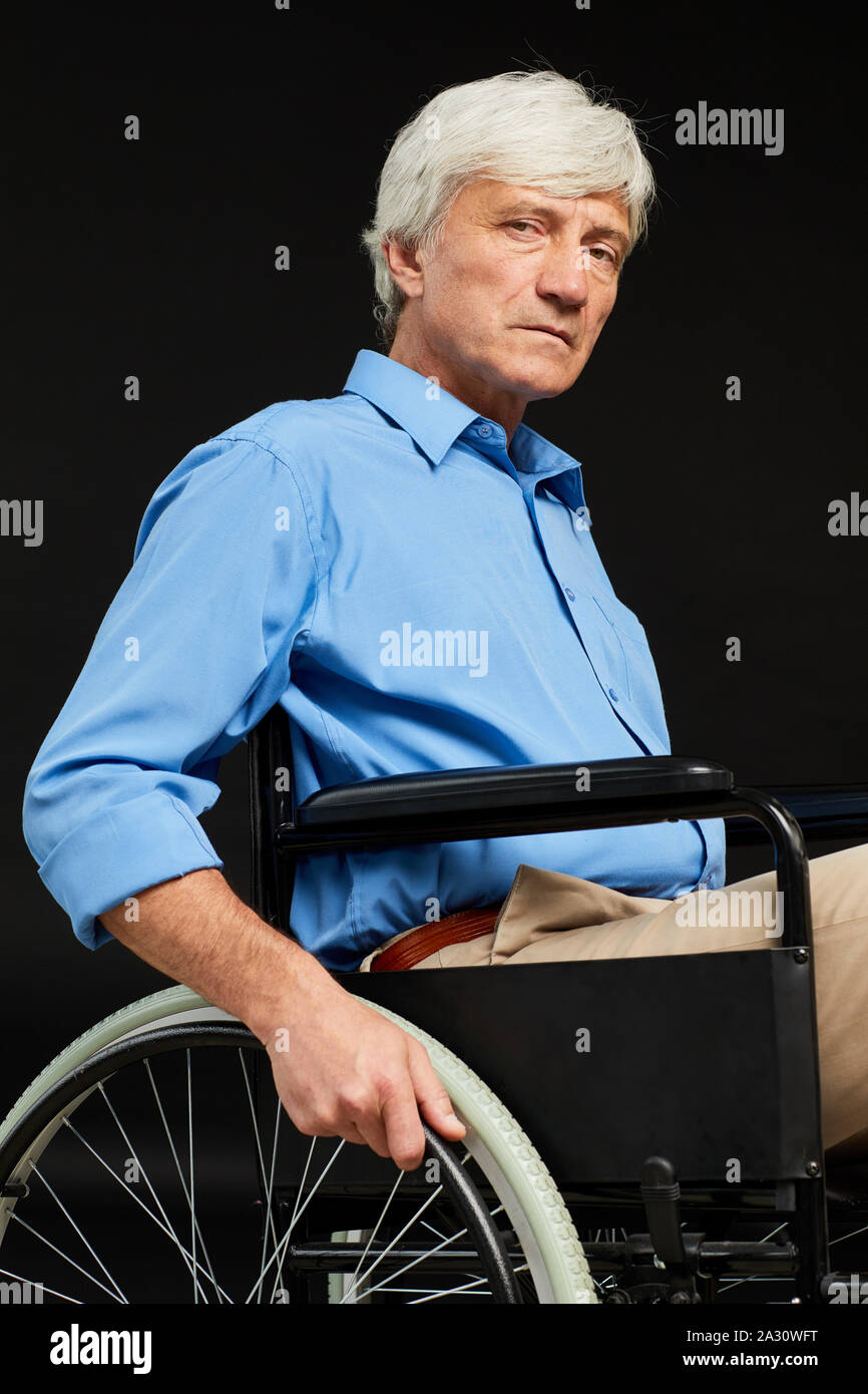 Portrait of senior man a une déficience il assis en fauteuil roulant et à la caméra à l'arrière-plan noir contre Banque D'Images