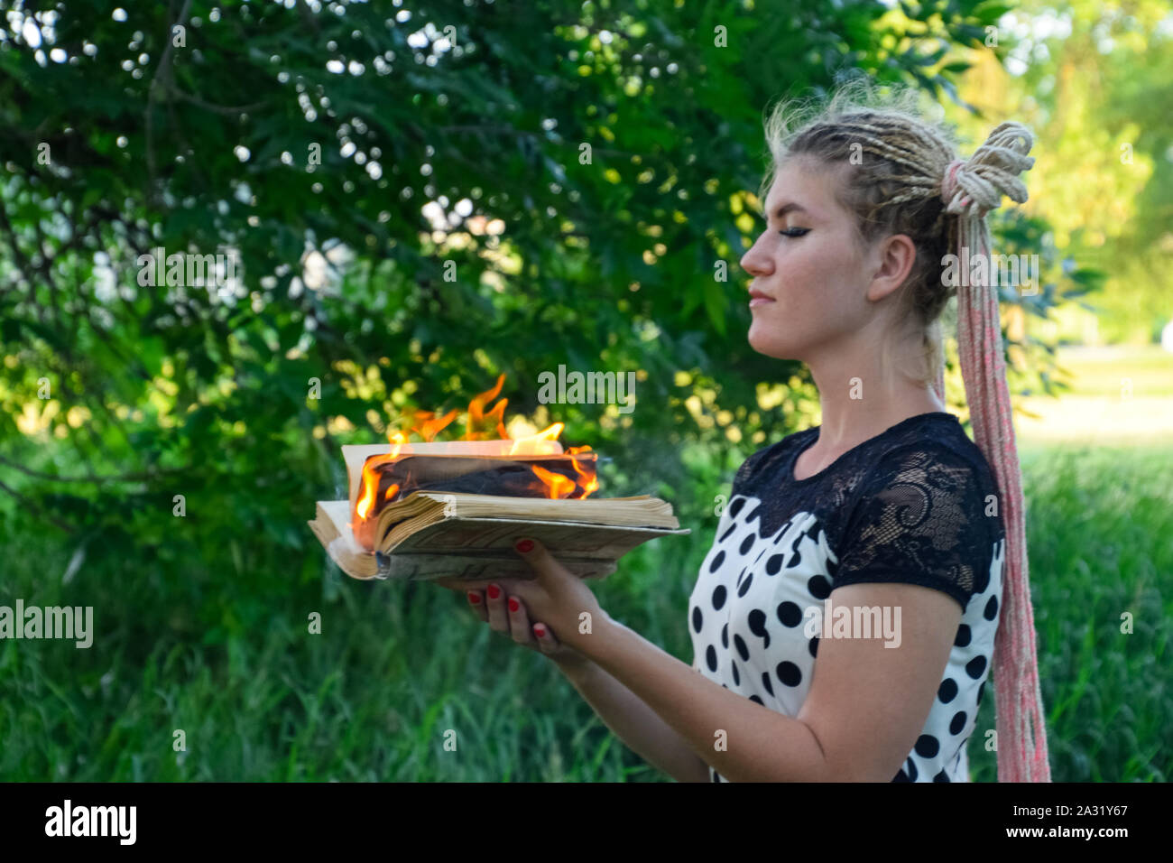 La jeune fille est titulaire d'un livre brûlant dans ses mains. Une jeune femme dans une forêt brûle un livre. Banque D'Images