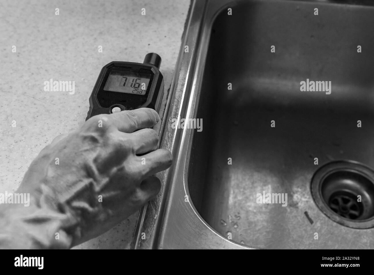 Un gros plan et vue monochrome sur la main d'un évaluateur de la qualité de l'environnement intérieur, prendre des lectures sur un compteur électronique par un évier de cuisine. Banque D'Images