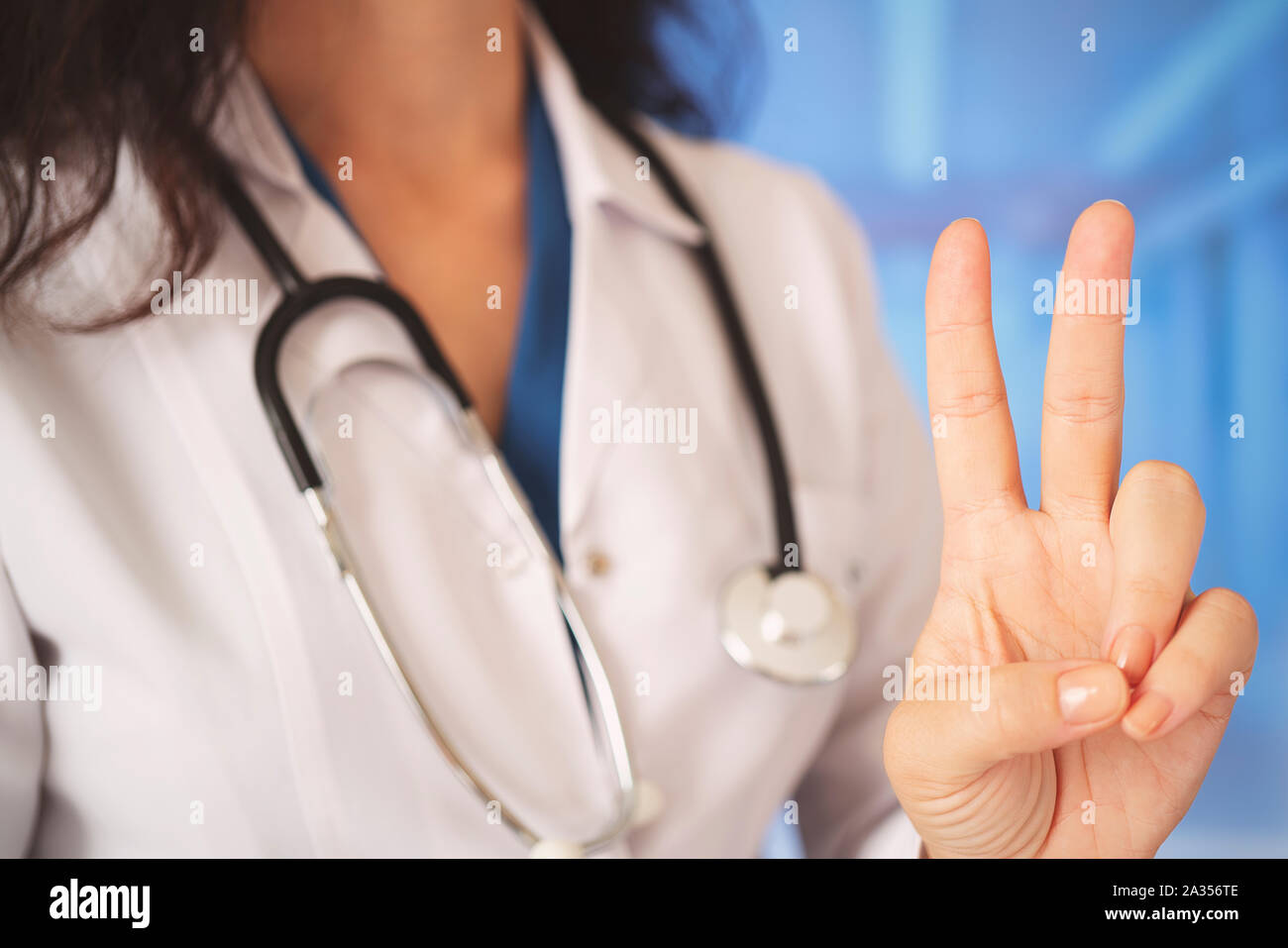 Belle jeune femme médecin à l'aide d'hôpital stéthoscope sur fond hall à la recherche de l'appareil photo montrant les doigts faisant signe de victoire. Numéro deux. Banque D'Images