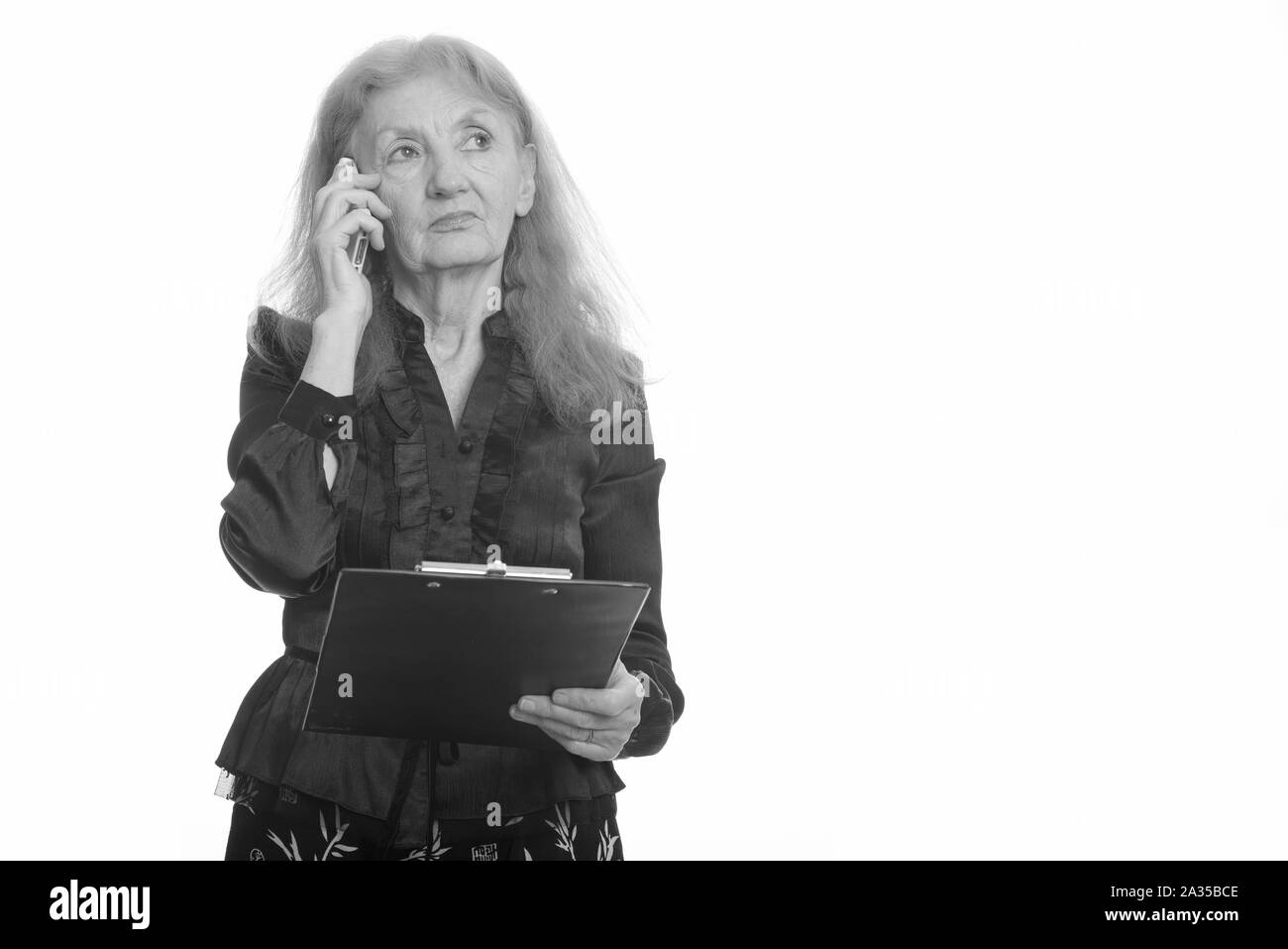 Studio shot of senior businesswoman holding clipboard while talking on mobile phone en pensant Banque D'Images
