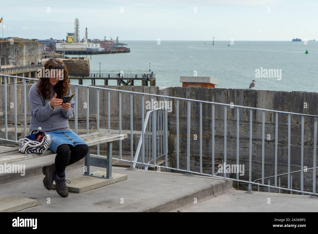 Une femme d'âge moyen assis sur un banc, à un point de vue de la mer à la recherche de son téléphone portable ou un téléphone cellulaire Banque D'Images