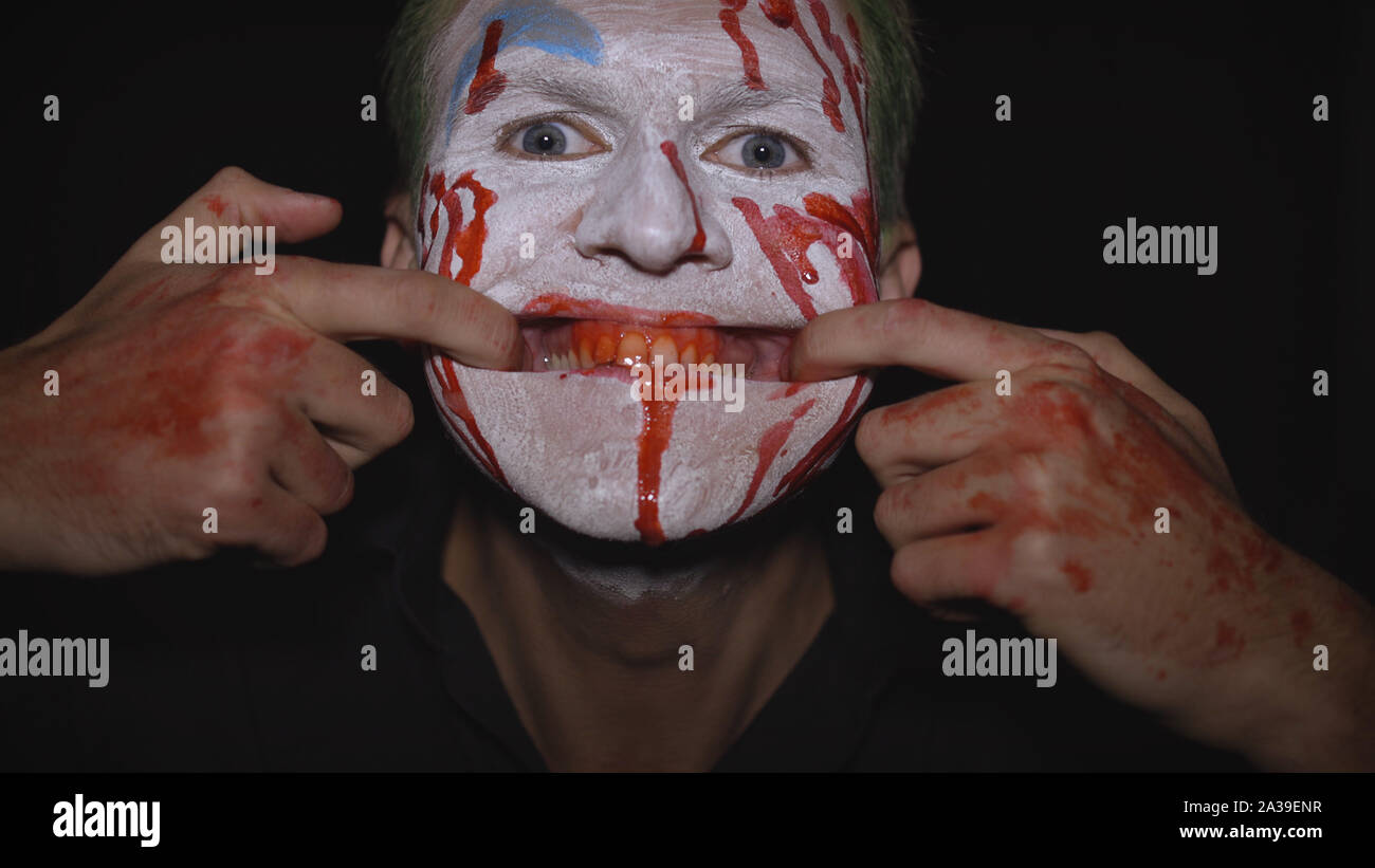 Clown Halloween Man Portrait. Close-Up De Fou, Le Mal De Clowns Face Avec  Le Sang. Maquillage Visage Blanc. Des Cheveux Verts. Sourire Effrayant Avec  Les Doigts. Modèle Attrayant En Costume D'halloween. Fond