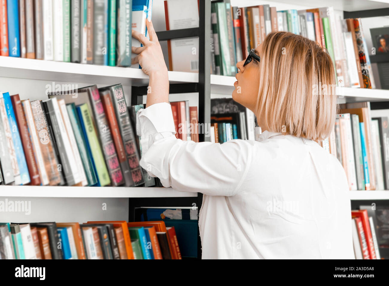 Jeune femme pensive attrayant à la main dessin un livre dans une librairie ou à la bibliothèque. La femme est sur le point de mire. Étagère avec les livres sont sur le premier plan et floue. Banque D'Images