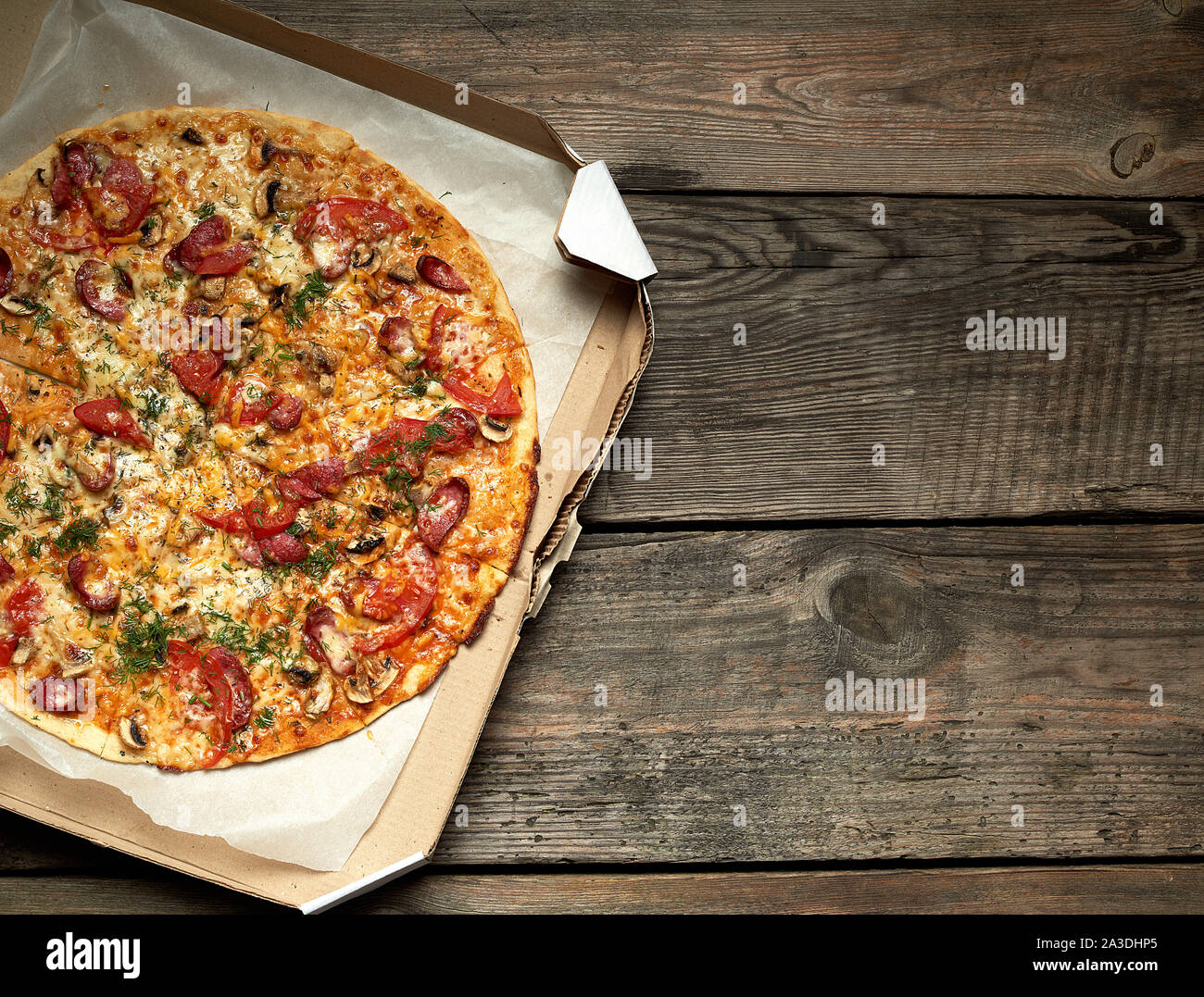 Ronde au four pizza avec saucisses fumées, champignons, tomates, fromage et de l'aneth dans une boîte en carton sur une table en bois, de l'alimentation est coupée en portions, t Banque D'Images