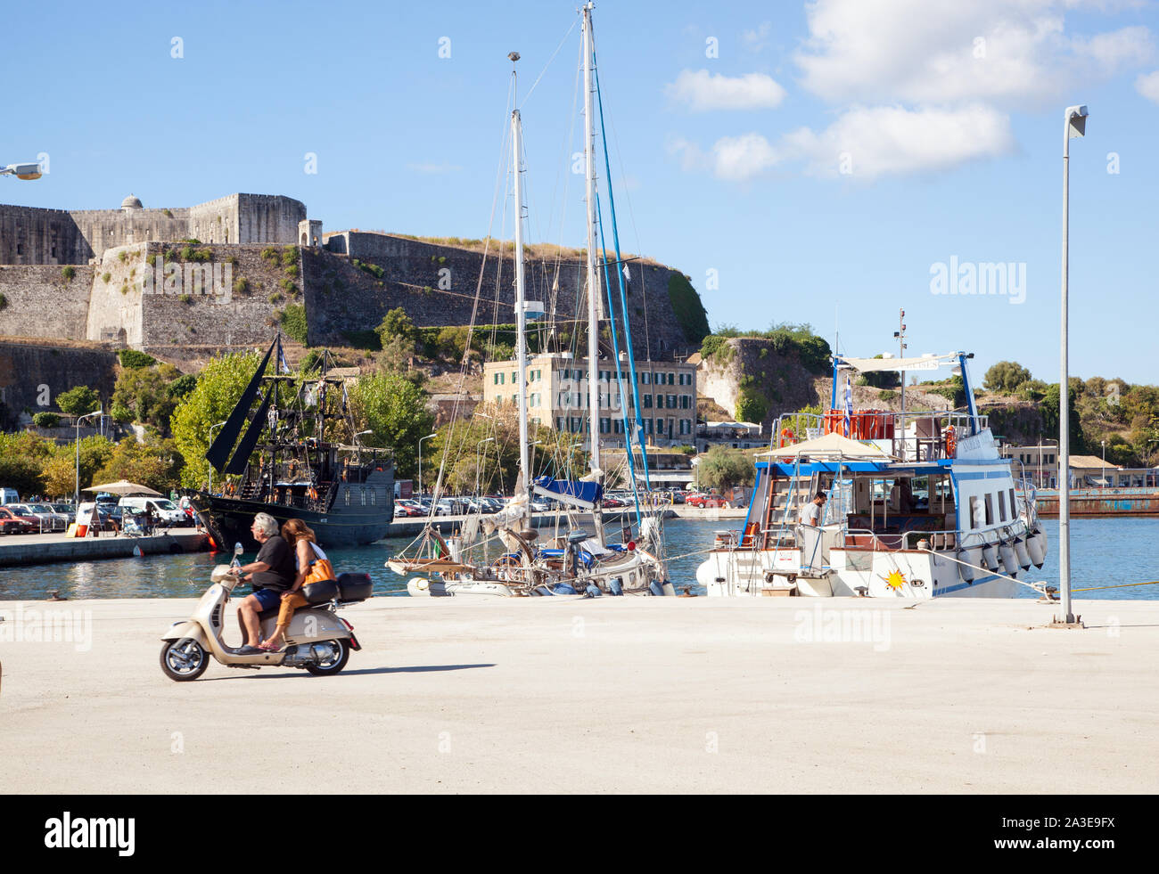 Man and Woman riding scooter de moteur sur le long le long du port de la ville grecque de Corfou Grèce avec le nouveau fort à l'arrière-plan Banque D'Images