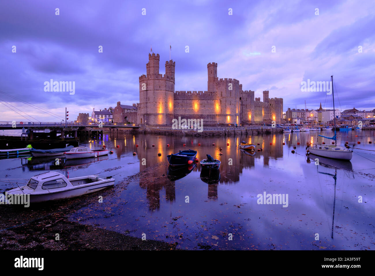 Château de Caernarfon éclairé au crépuscule ce qui traduit en baie avec bateaux dans Port Moody ld'purple ciel nuageux dans le Nord du Pays de Galles Banque D'Images