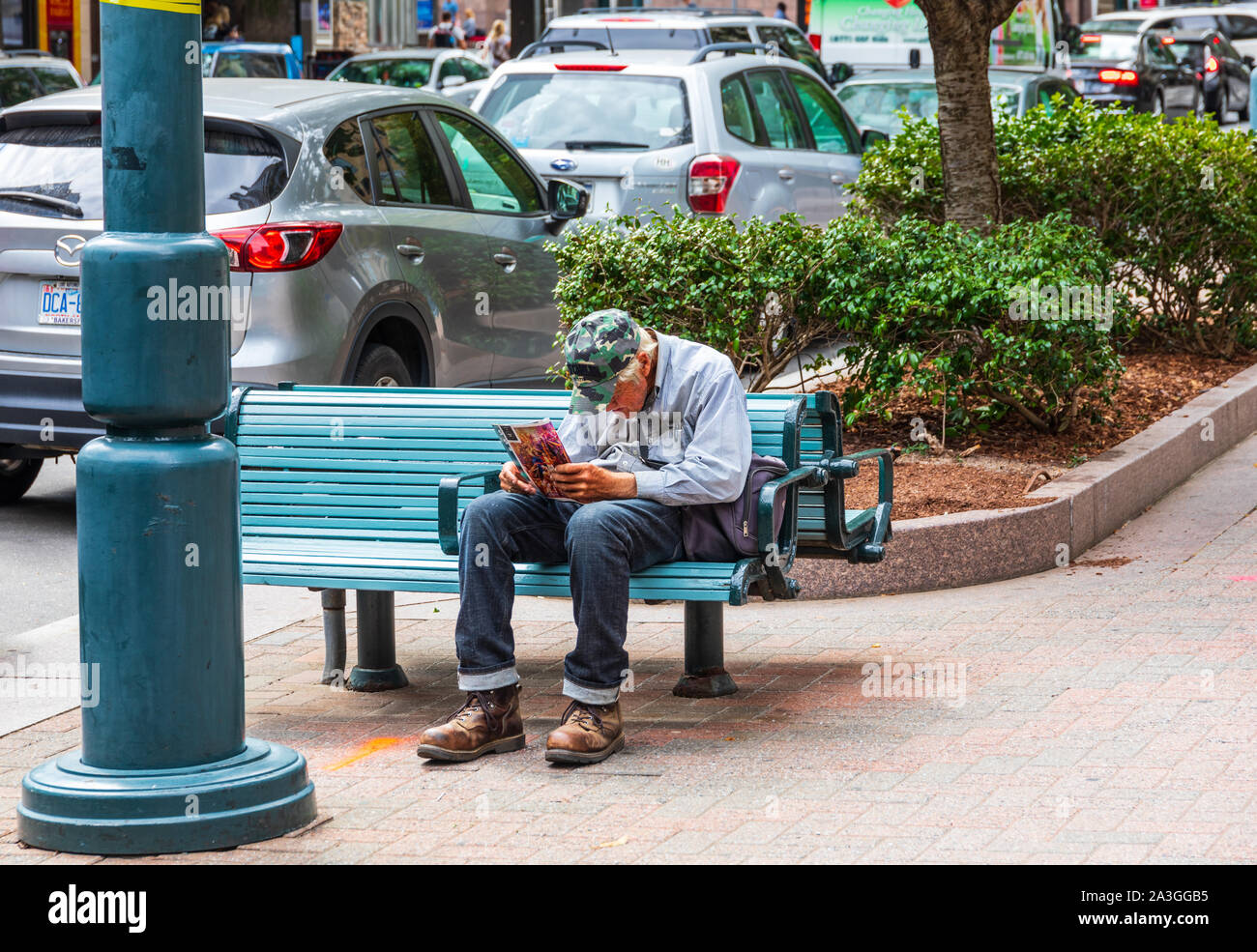 CHARLOTTE, NC, USA-6 oct 2019 : Un homme âgé avec siège arrière voûtée sur un banc sur Tryon Street, la lecture d'un magazine. Banque D'Images