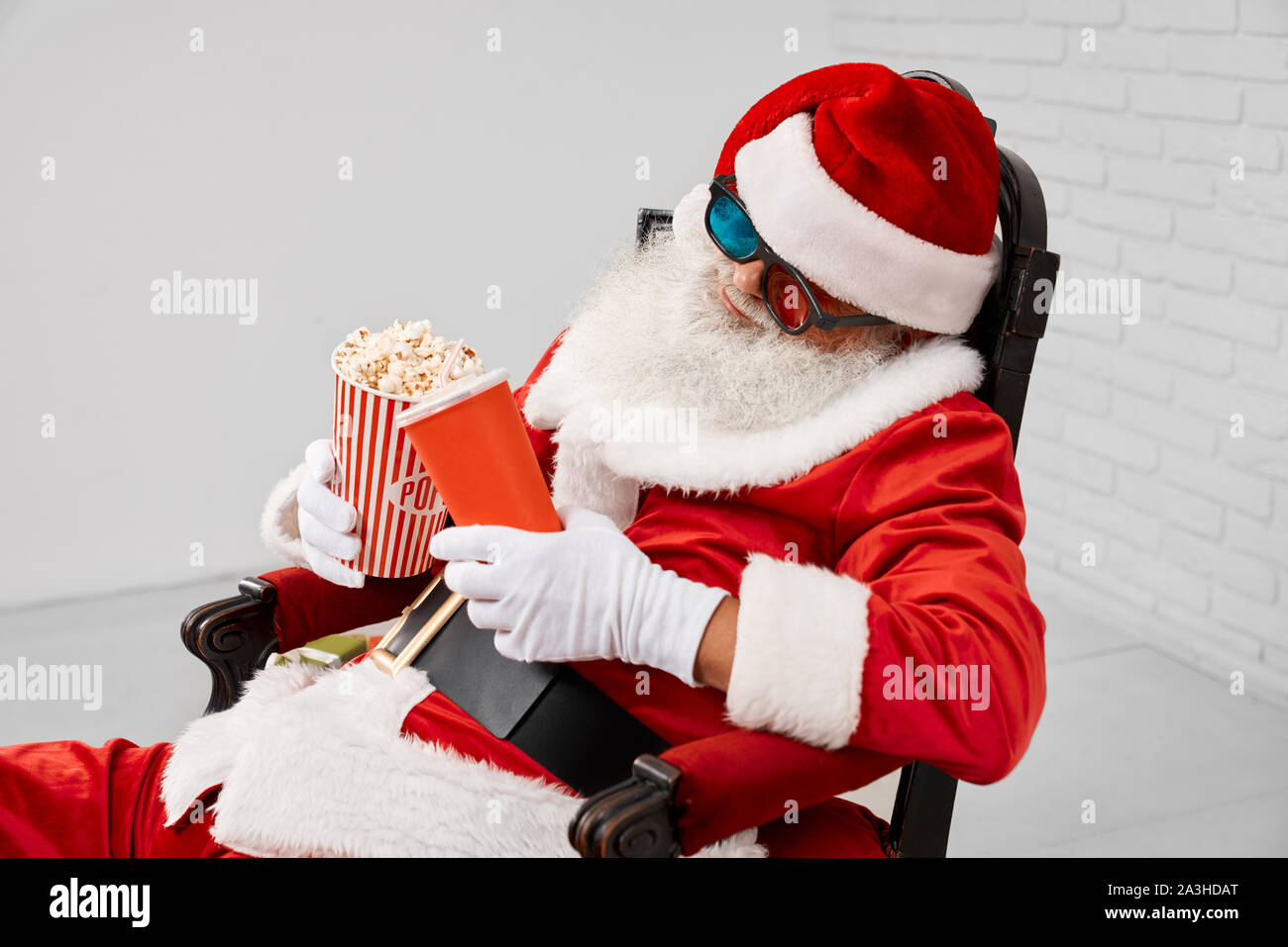 Saint Nicolas à dormir dans les lunettes 3D, red hat et gants blancs se détendre dans un fauteuil tout en maintenant seau de popcorn et cola en mains. Le temps de Noël et des traditions nationales. Banque D'Images