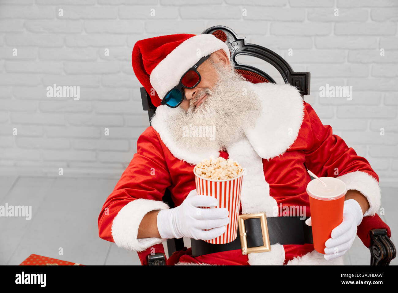 Le caractère traditionnel de Noël fatigué en costume rouge dormir dans un fauteuil confortable avec pop-corn et de coca dans les mains. Le Père Noël à maturité en tenant sieste avant de donner des cadeaux à des enfants heureux. Banque D'Images