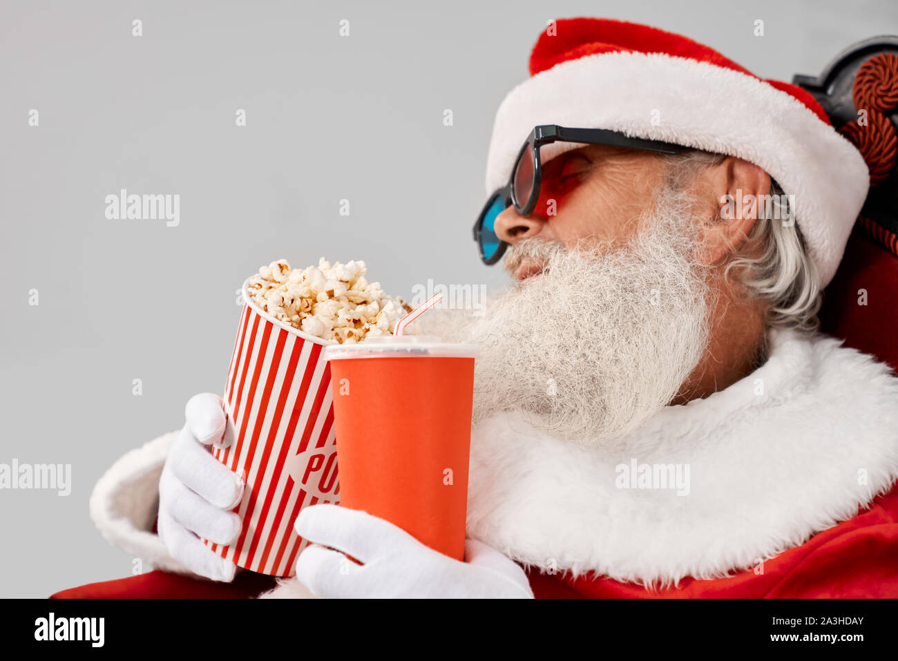 Vue de côté de Santa Claus dans red hat, gants blancs et 3D sans lunettes s'endormir en regardant film avec pop-corn et de coca dans les mains. Man costume noël dormir dans un fauteuil Banque D'Images
