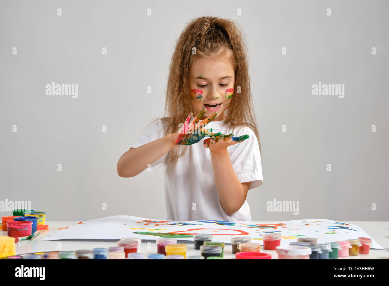 Petite fille en t-shirt blanc assis à table avec les peintures et whatman sur elle, posant avec un visage peint et les mains. Isolé sur blanc. Medium close-up. Banque D'Images