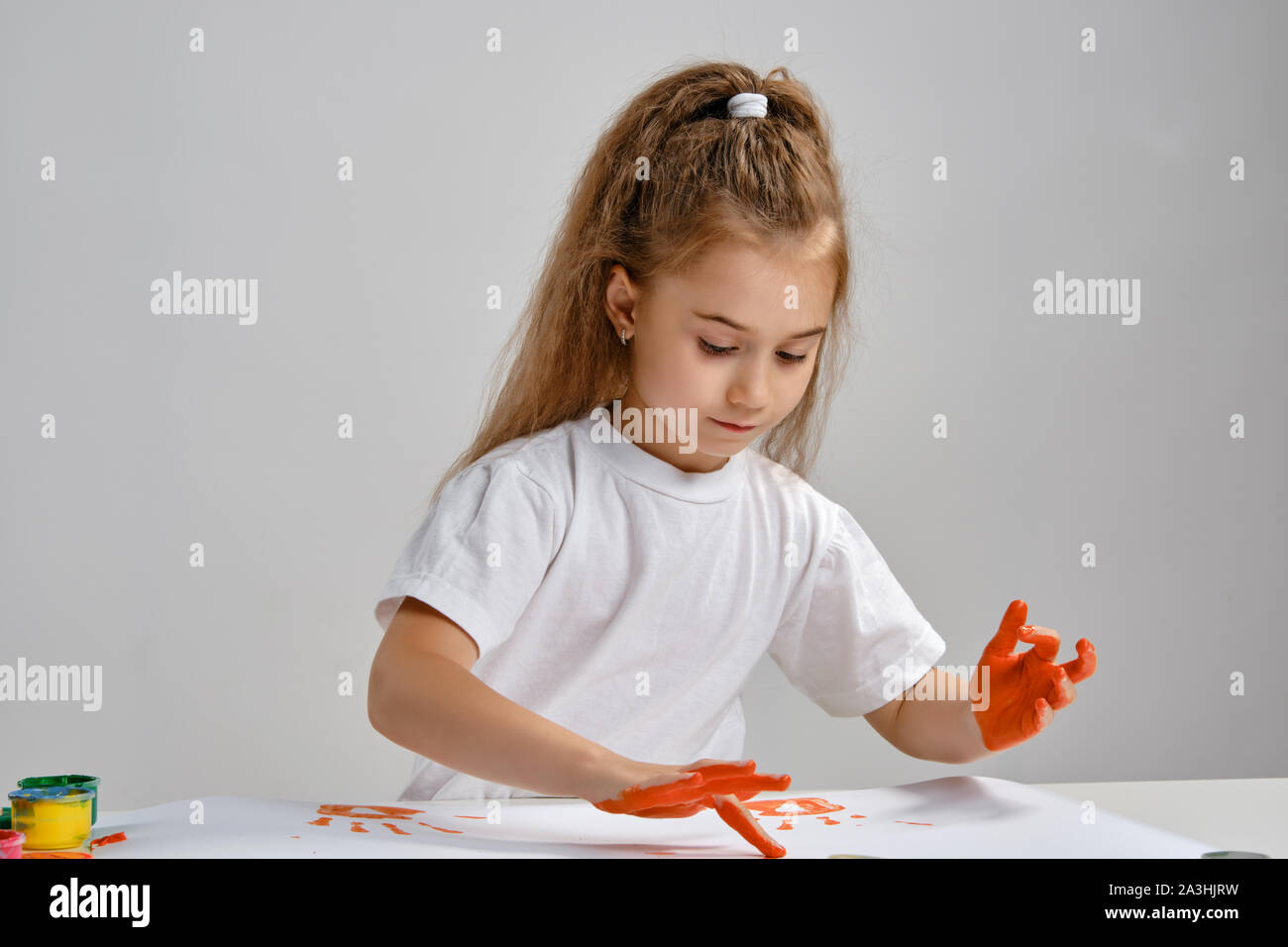 Petite fille en t-shirt blanc assis à table avec des peintures colorées et Whatman, peinture sur elle avec ses mains. Isolé sur blanc. Medium close-up. Banque D'Images