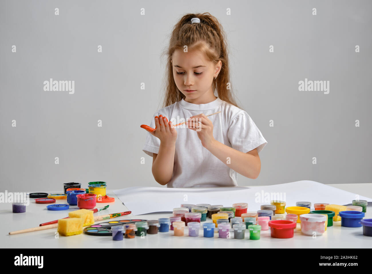 Petite fille en t-shirt blanc assis à table avec des peintures colorées et whatman sur elle, la peinture ses mains. Isolé sur blanc. Medium close-up. Banque D'Images