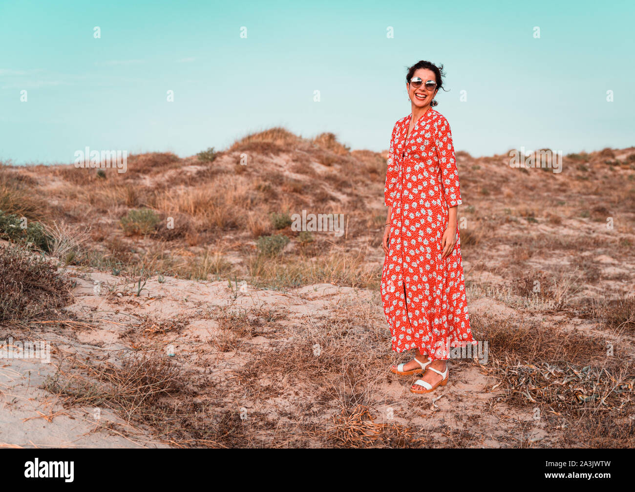 Beautiful smiling woman in red dress et round sunglasses smiling dans des dunes de sable blanc de la Plage de Oliva au coucher du soleil. Femme sur son 30. Suitab Banque D'Images