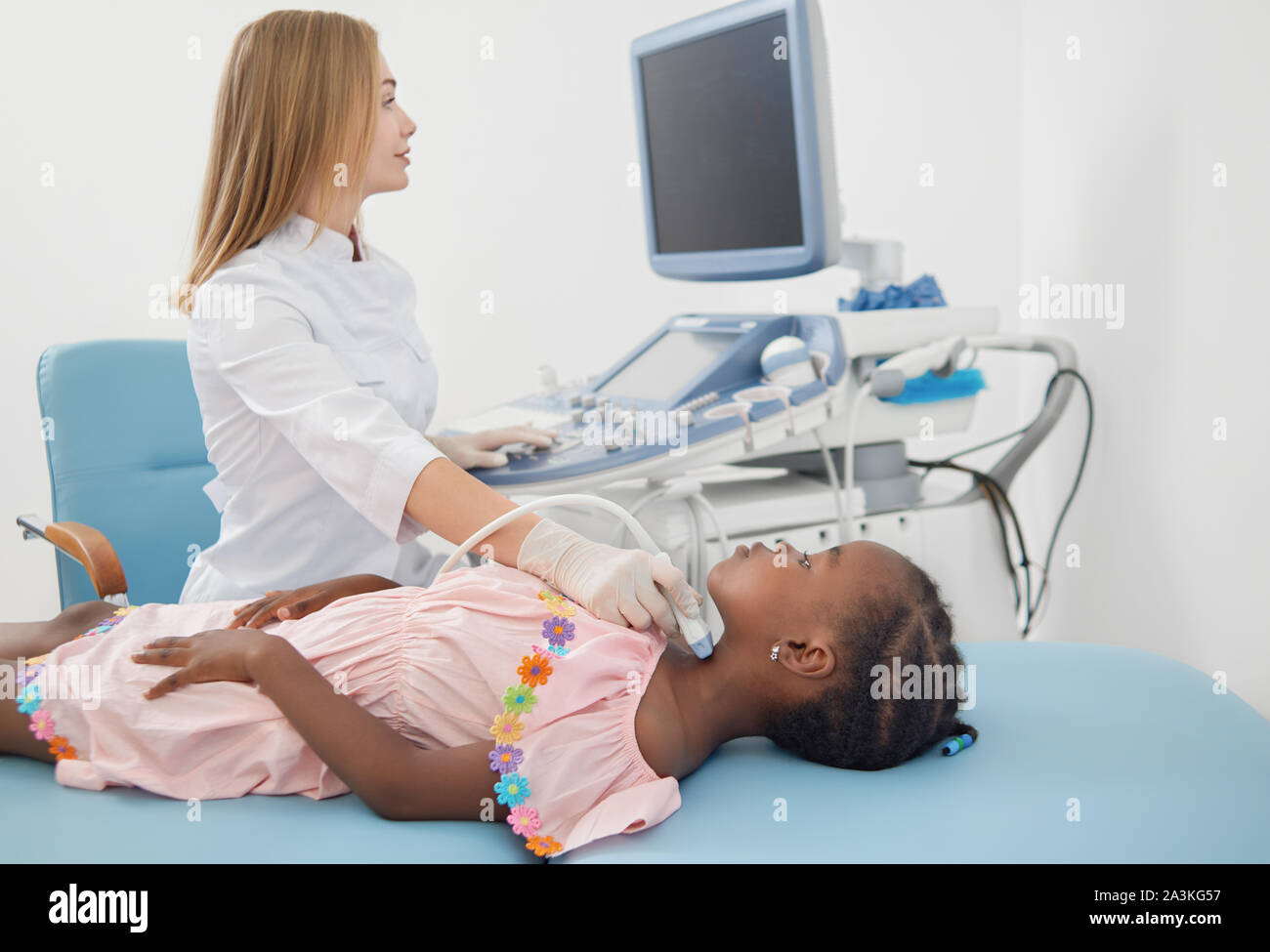 Joli enfant afro lying on bed, au cours de l'échographie de la thyroïde. Petite fille, vêtu en robe rose, courtoise suit les instructions de la femme médecin avec sonde échographique dans la main et la numérisation de son cou. Banque D'Images