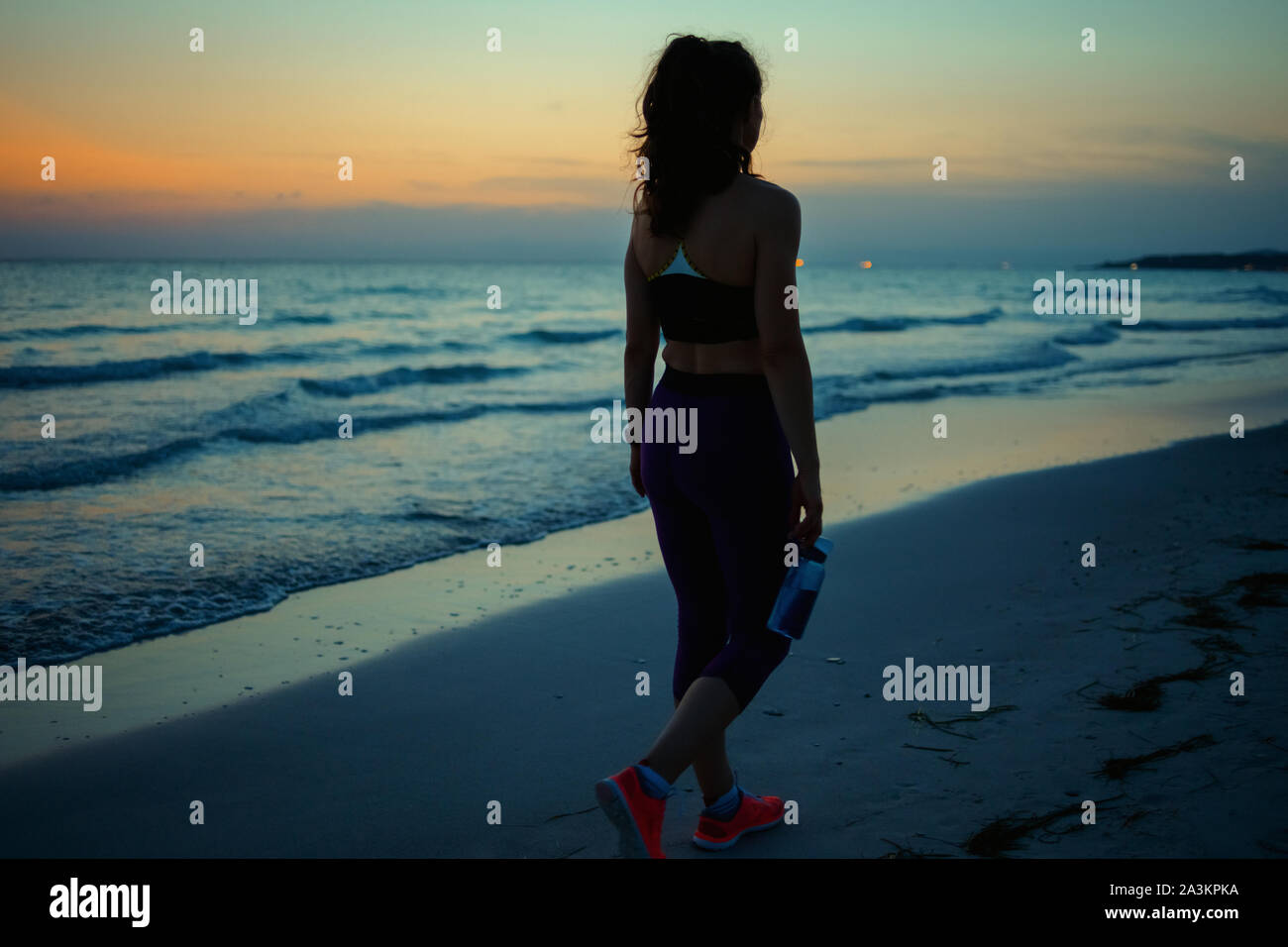 Vus de derrière, les sports fitness femme en vêtements de fitness avec une bouteille d'eau de marcher après entraînement sur la rive de l'océan dans la soirée. Banque D'Images