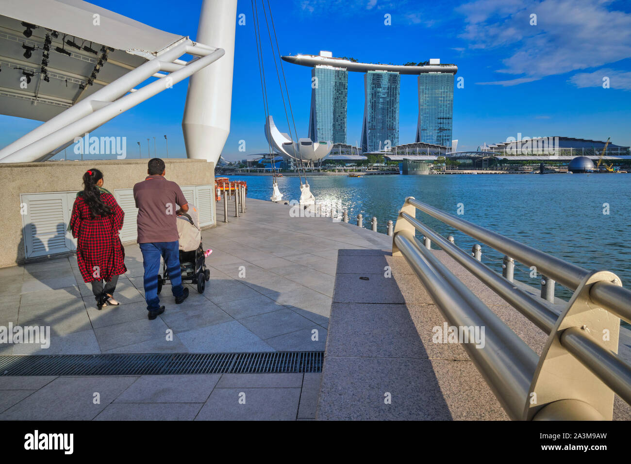 Vue depuis l'Esplanade de Singapour à travers Marina Bay en direction de Marina Bay Sands Hotel et musée ArtScience, avec un couple avec une poussette de marche Banque D'Images