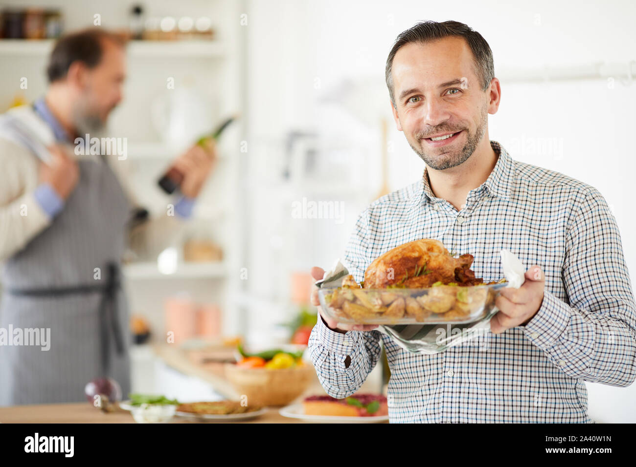 Portrait of young man holding plat de poulet rôti et smiling at camera tandis que d'autres homme de préparer des aliments à l'arrière-plan Banque D'Images