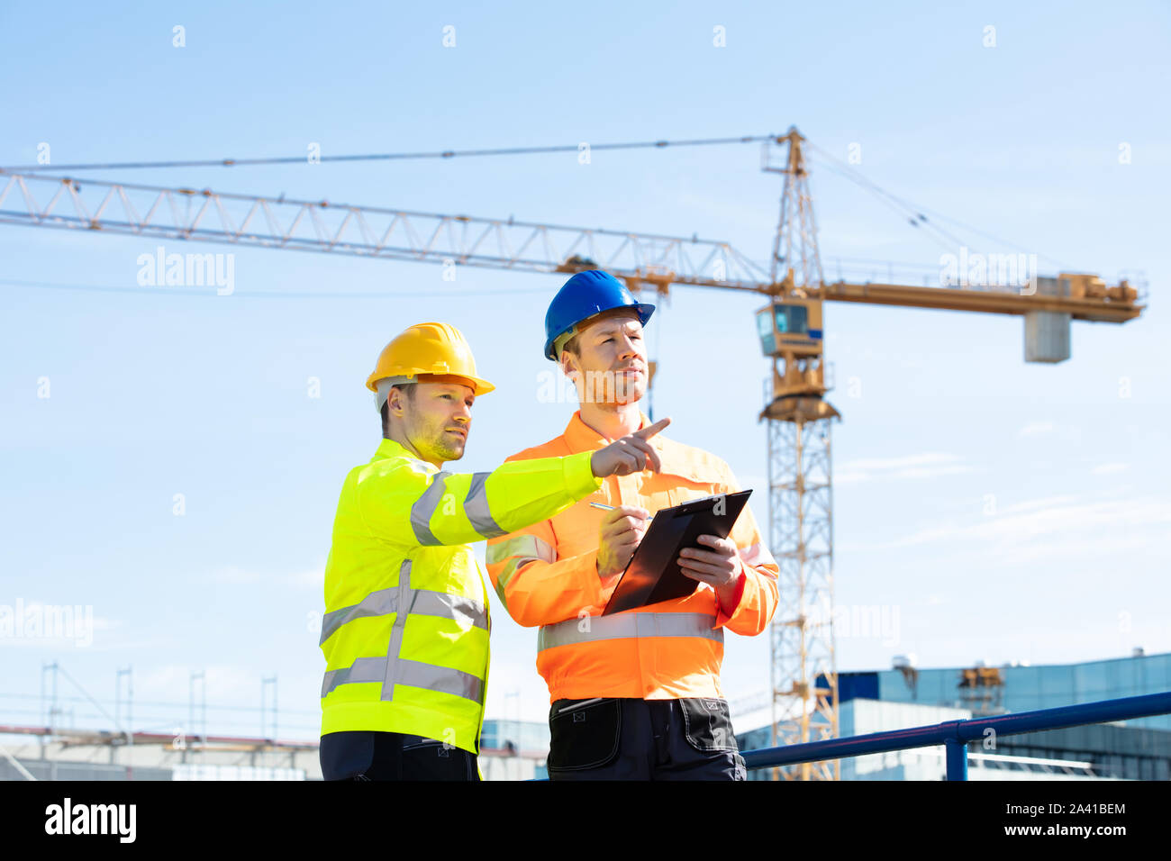 Deux jeunes ingénieurs masculins debout en face de la grue mécanique travaillant sur le chantier de construction Banque D'Images