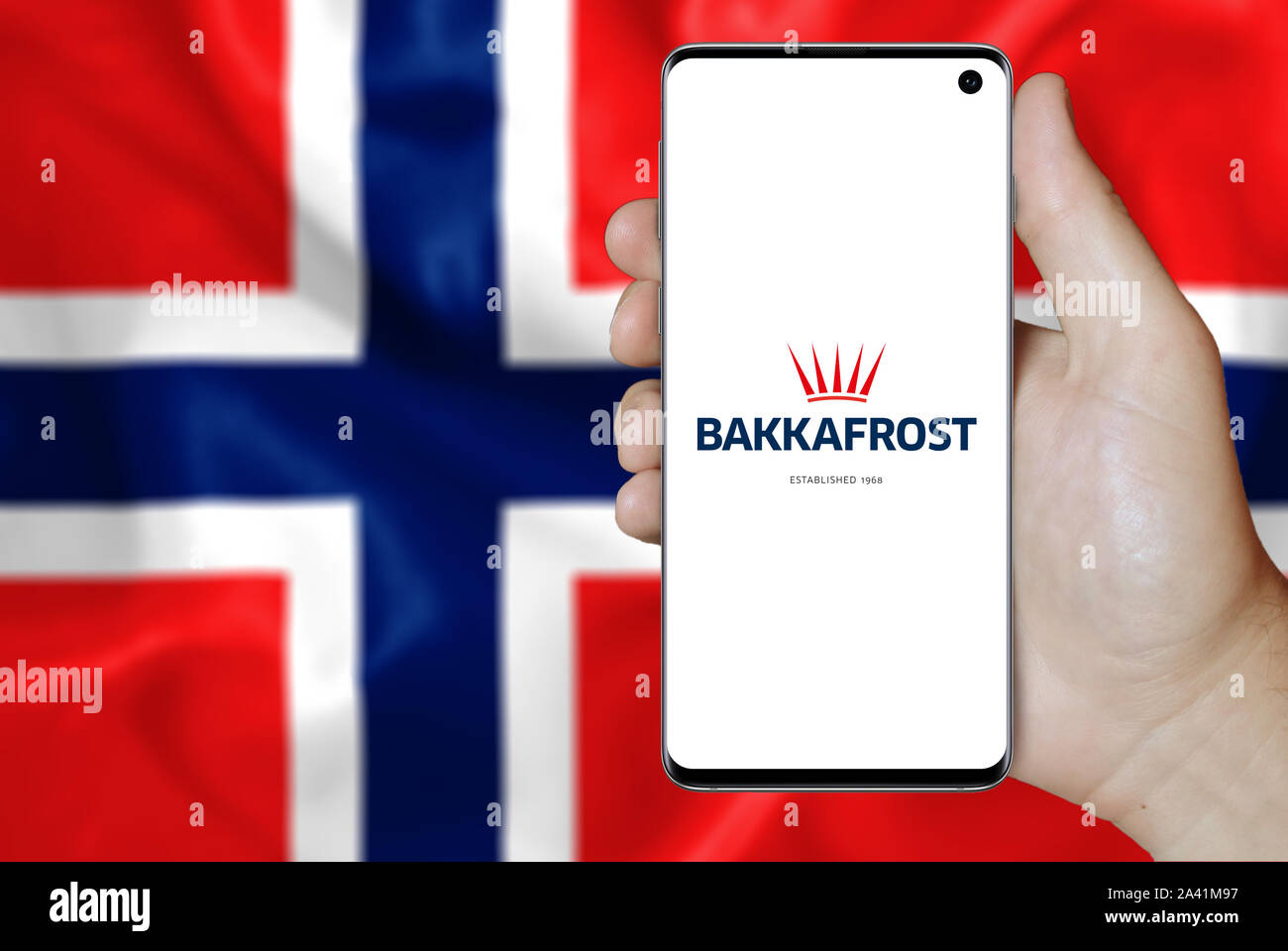 Un homme est titulaire d'un téléphone d'afficher le logo de société Bakkafrost inscrit sur la Bourse d'Oslo. Pavillon norvégien arrière-plan. Credit : PIXDUCE Banque D'Images