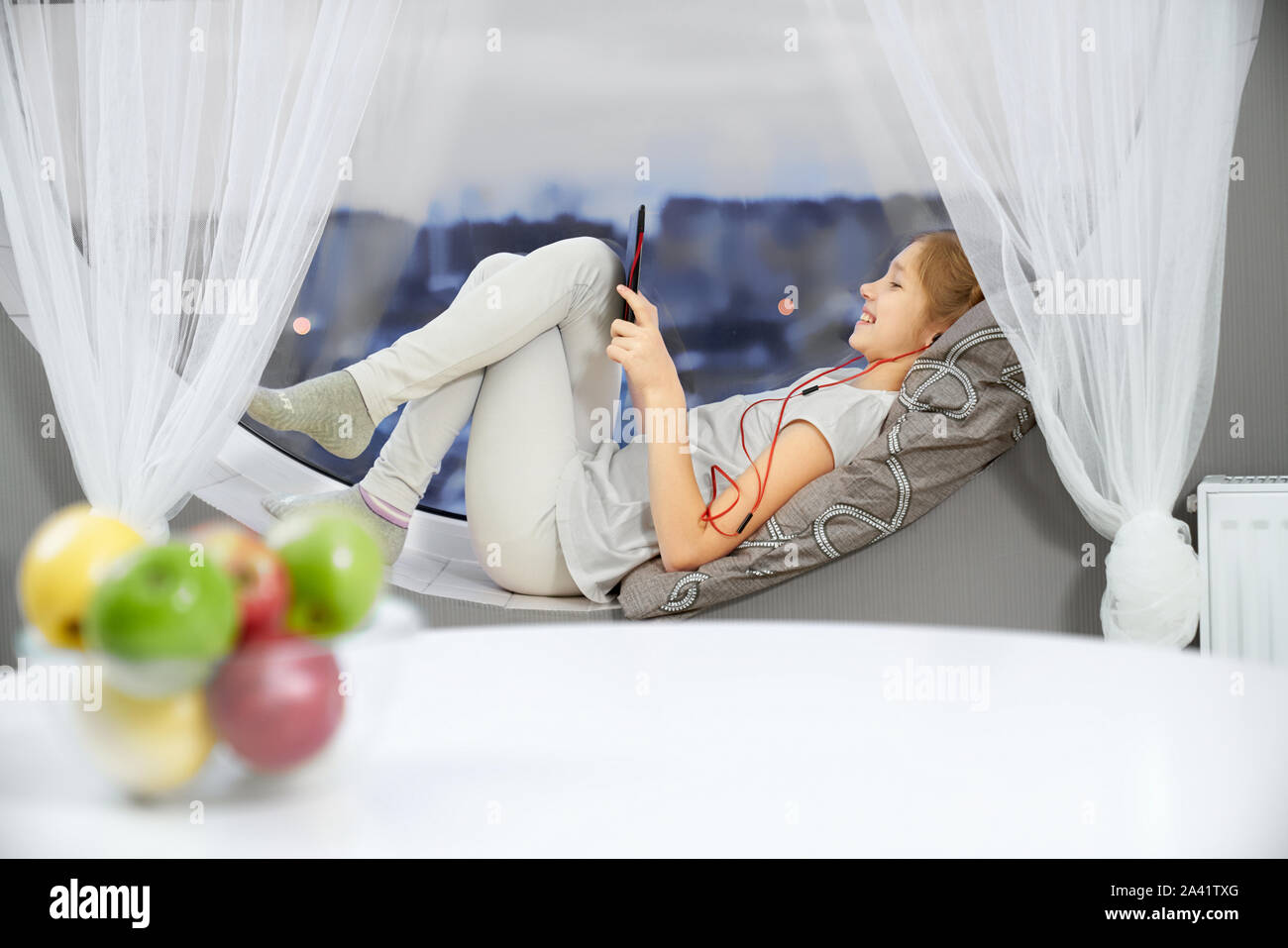 Vue latérale du rire adolescent détendue, allongé sur un canapé, écouter la musique et regarder des vidéos sur votre tablette. Girl resting et sirfing dans internet à la maison. Les pommes en restant sur la plaque de table. Concept de reste. Banque D'Images