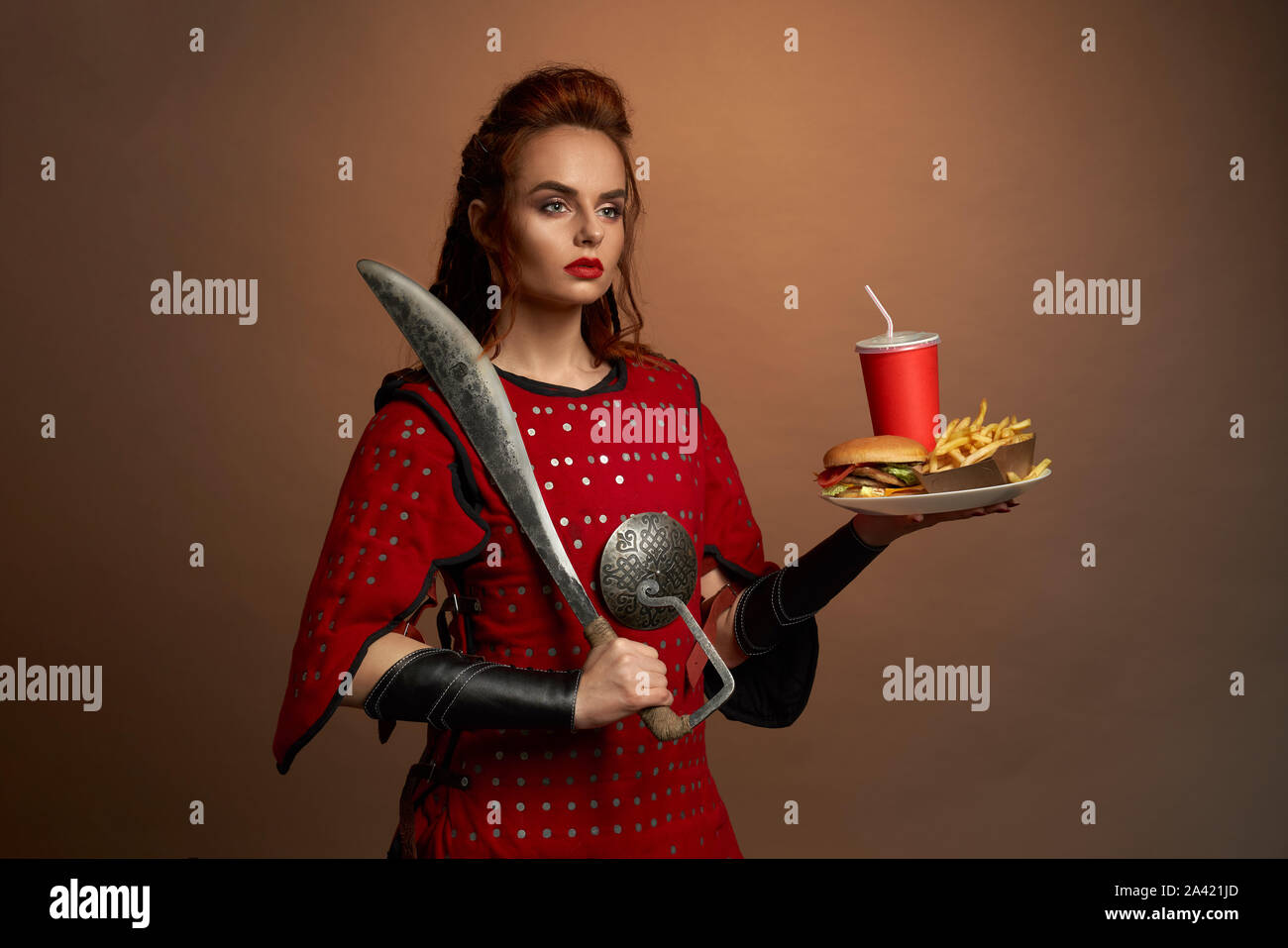 Belle femme fighter en armure rouge garder grand couteau et la plaque avec la restauration rapide en studio. Femme déterminée à côté et posant avec un hamburger et une pomme de terre. Concept de l'alimentation des héros. Banque D'Images