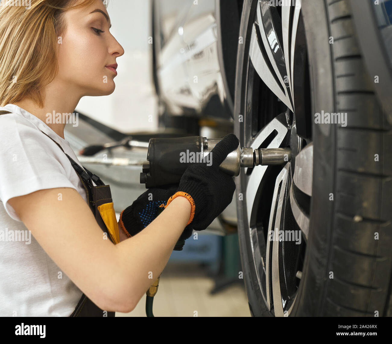 Vue latérale des belles femmes de pneu changer de mécanicien automobile en atelier. Blonde en chemise blanche, combinaison et gants de protection travaillant dans le service automobile. Concept d'entretien et de réparation. Banque D'Images