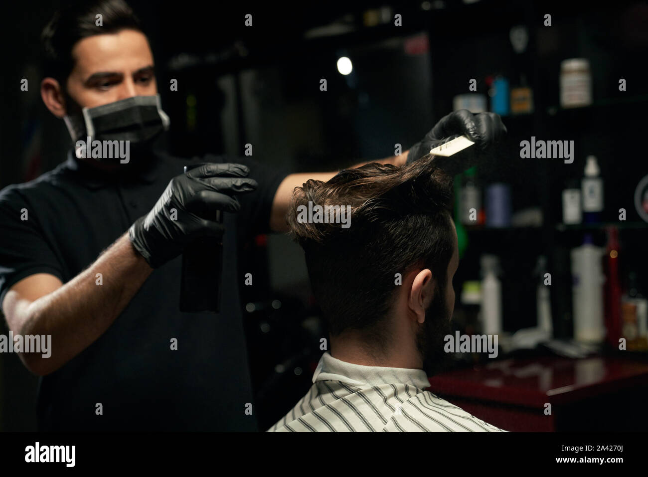 Salon de coiffure professionnel hommes habillés en chemise noire, le port de masque sur la bouche et sur les mains des gants est en train de faire de la coiffure pour homme unshaved que couvert de cape rayée assis dans un fauteuil au salon de coiffure Banque D'Images