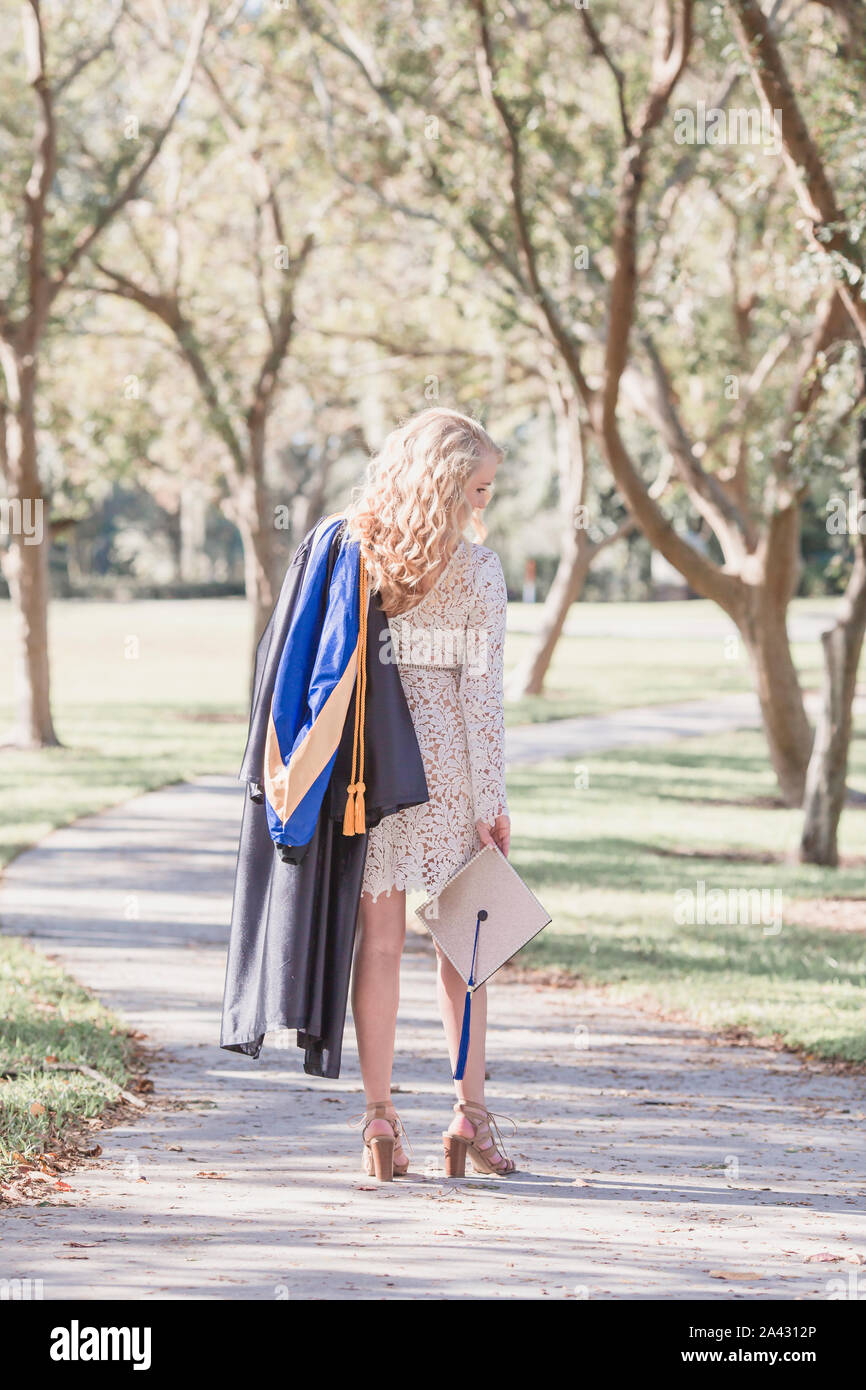 Femme blonde long cheveux bouclés détient graduation cap and gown dans chemin Banque D'Images