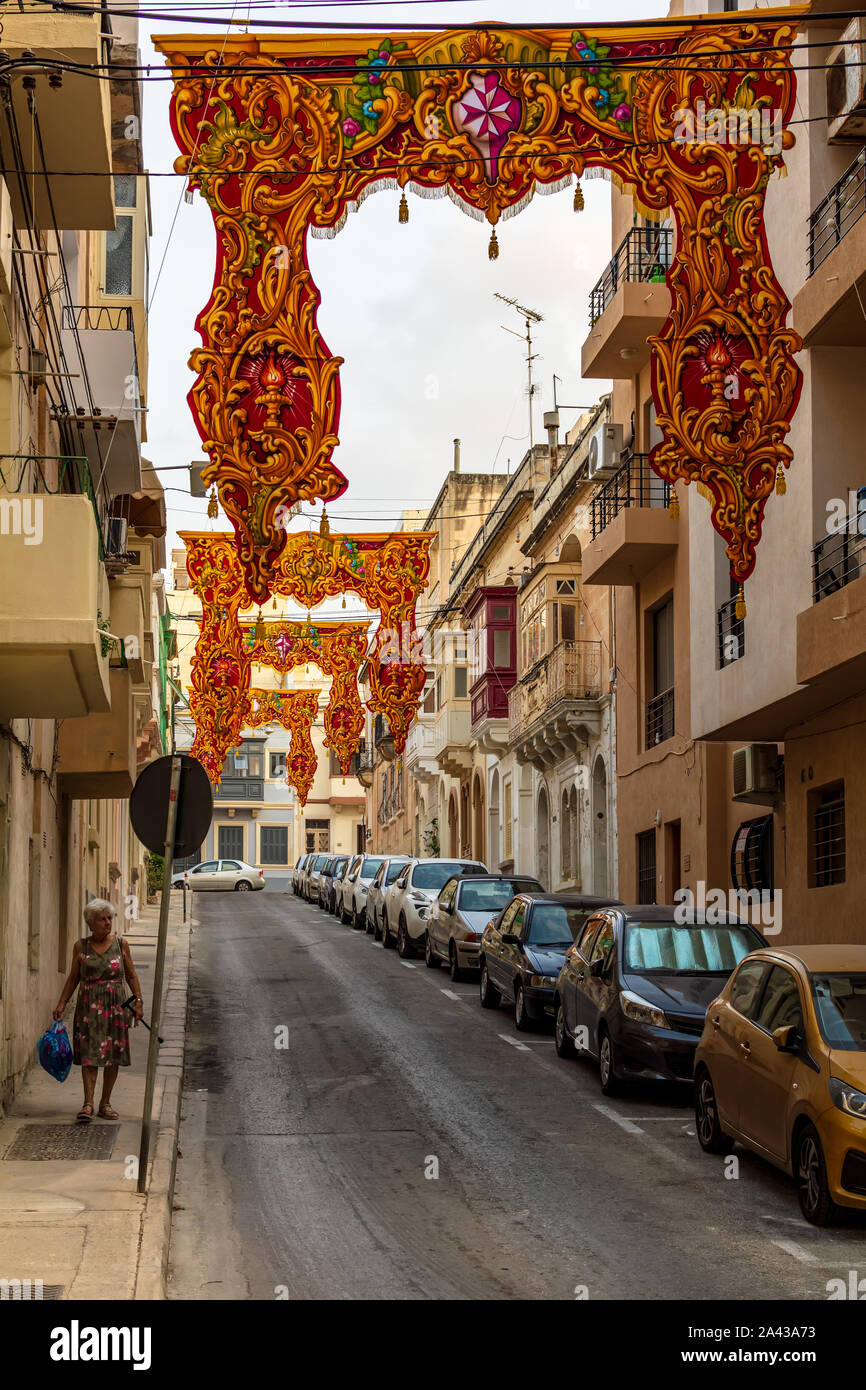 Vieille femme marchant dans la rue à Sliema décoré avec des ornements religieux rouge bannières pour Fête du village (festa) de Saint Grégoire le Grand. Banque D'Images