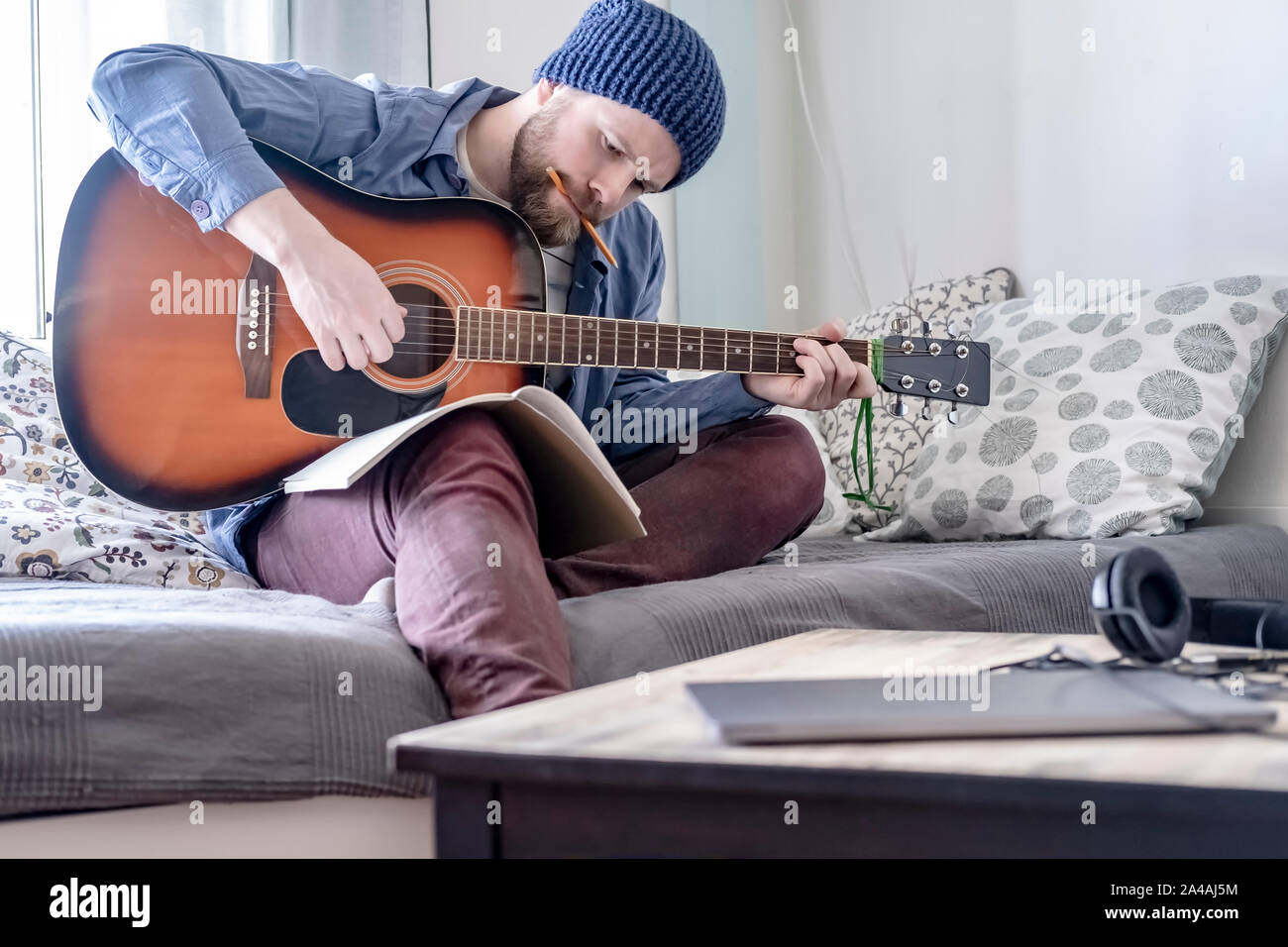 L'accent jeune compositeur écrit la musique joue sur une guitare acoustique tout en regardant un carnet et un crayon dans la bouche, assis sur un canapé dans un co Banque D'Images