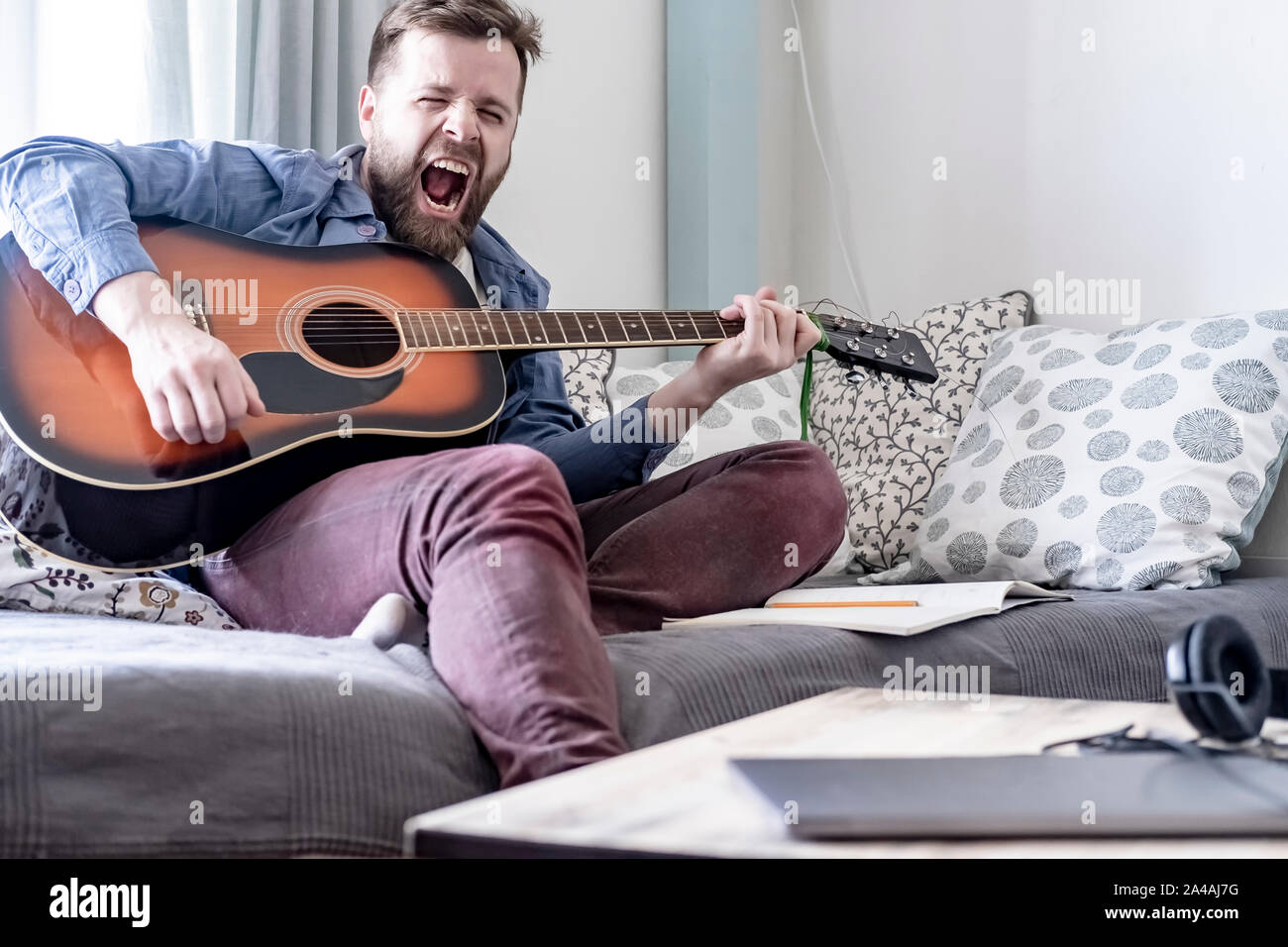 Un jeune, beau mâle compositeur joue une guitare acoustique et un chant haut et fort de sa propre composition, assis sur un canapé, dans une maison confortable envi Banque D'Images