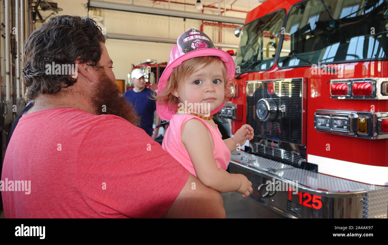 A cute baby girl wearing a helmet fire rose sourit à l'appareil photo de son père au cours d'armes du poste d'incendie portes ouvertes. Banque D'Images