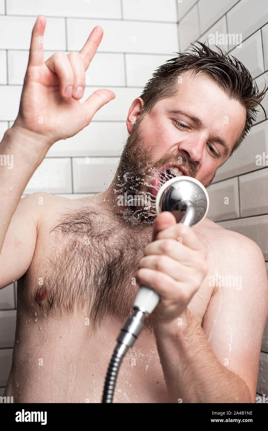 Un homme barbu chante dans la salle de bains, à l'aide d'une douche à l'eau courante au lieu d'un microphone et montrant rock n roll signer ou donner le diable cornes Banque D'Images