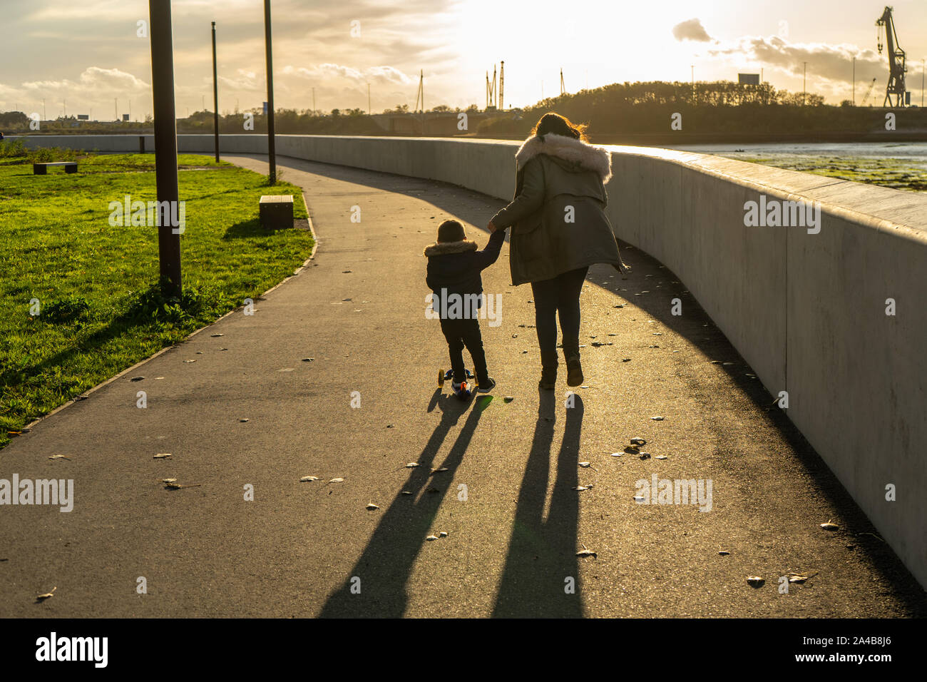 Une mère marche sur le trottoir avec son jeune fils de la trottinette à côté d'elle lui tenant la main au coucher du soleil Banque D'Images