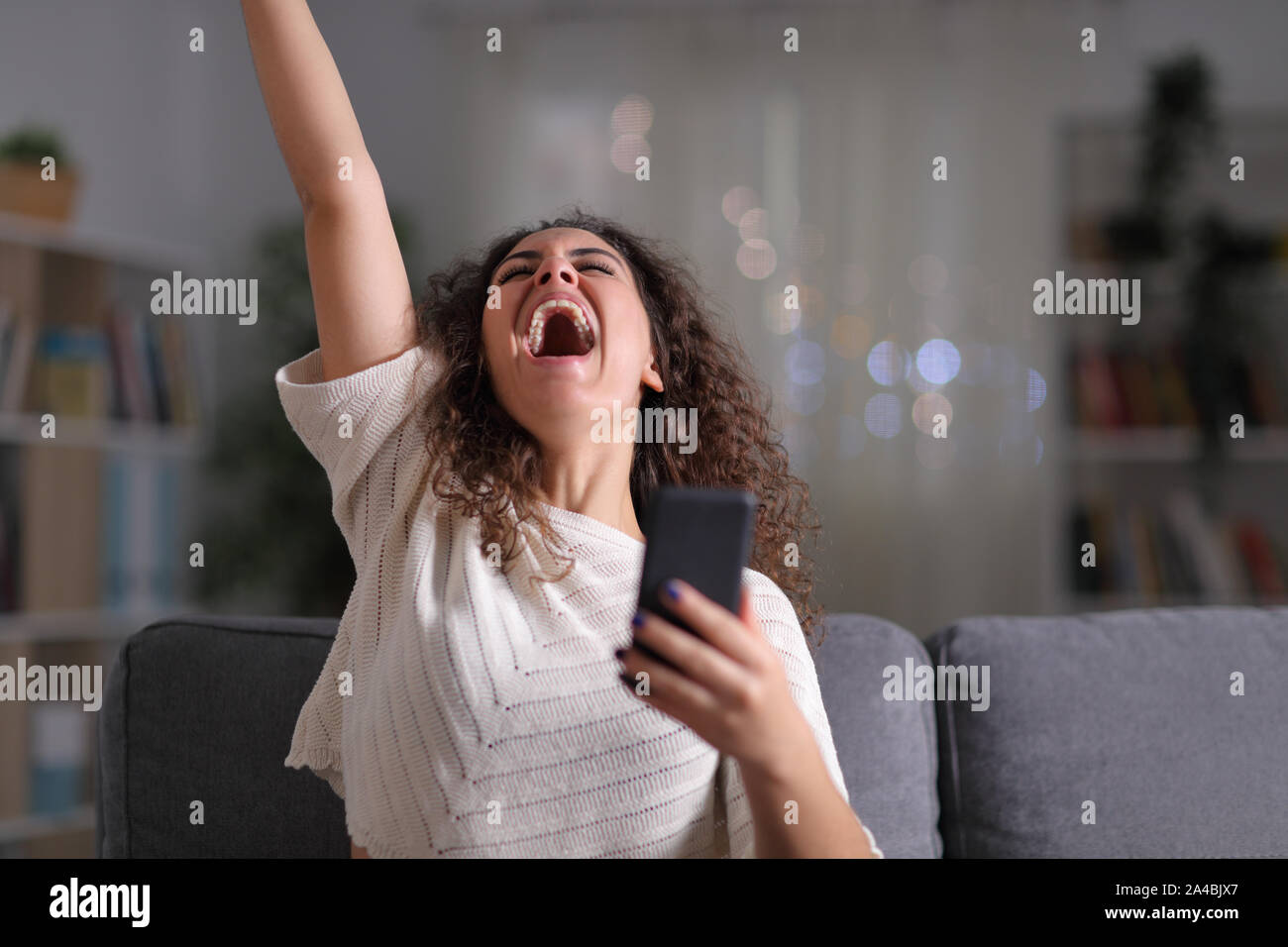 Excited woman celebrating success with assis sur un canapé dans la nuit à la maison Banque D'Images
