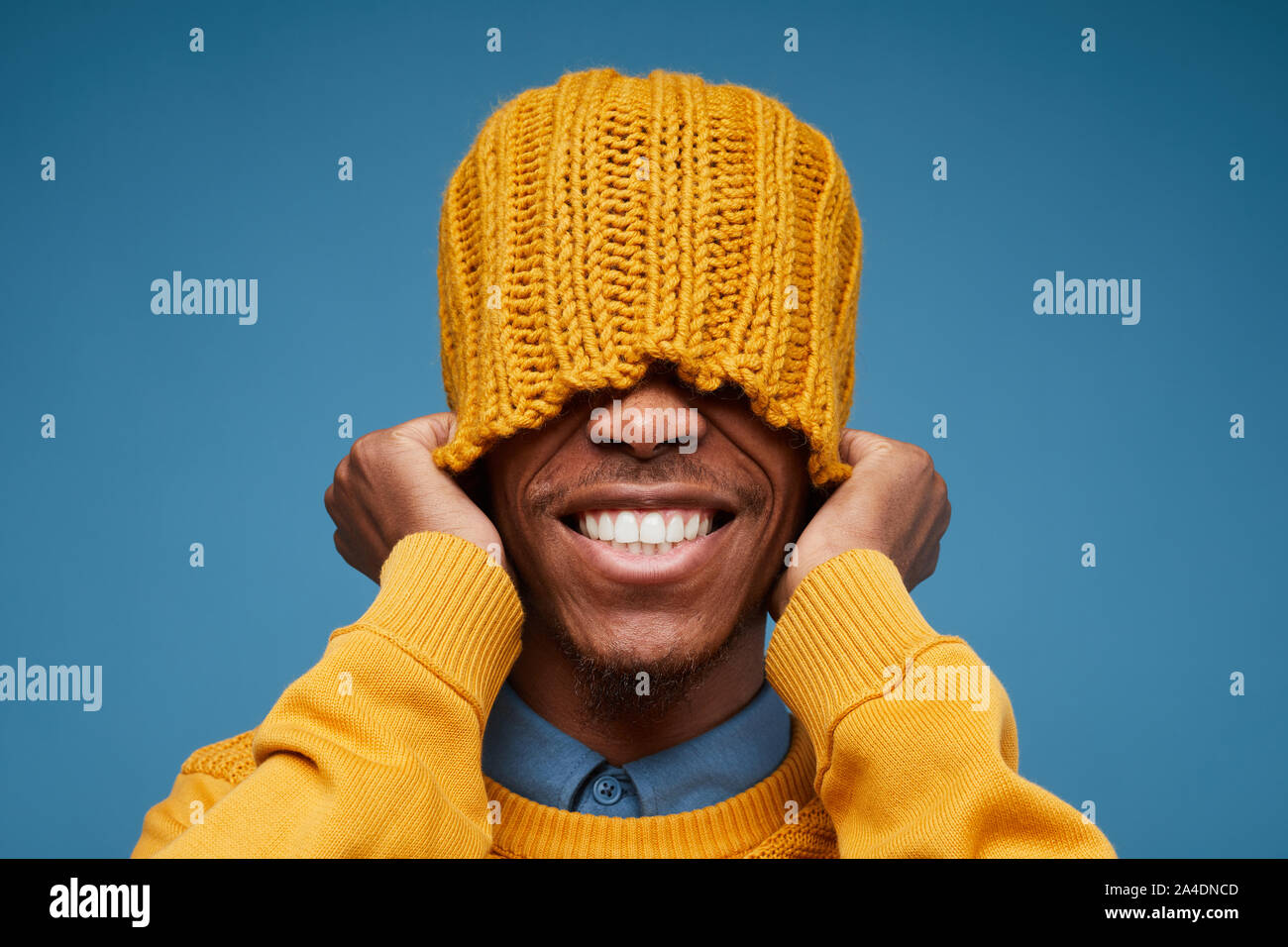 Portrait de vives émotions African man smiling at camera tout en tirant sur le jaune Knit hat posing contre fond bleu, copy space Banque D'Images