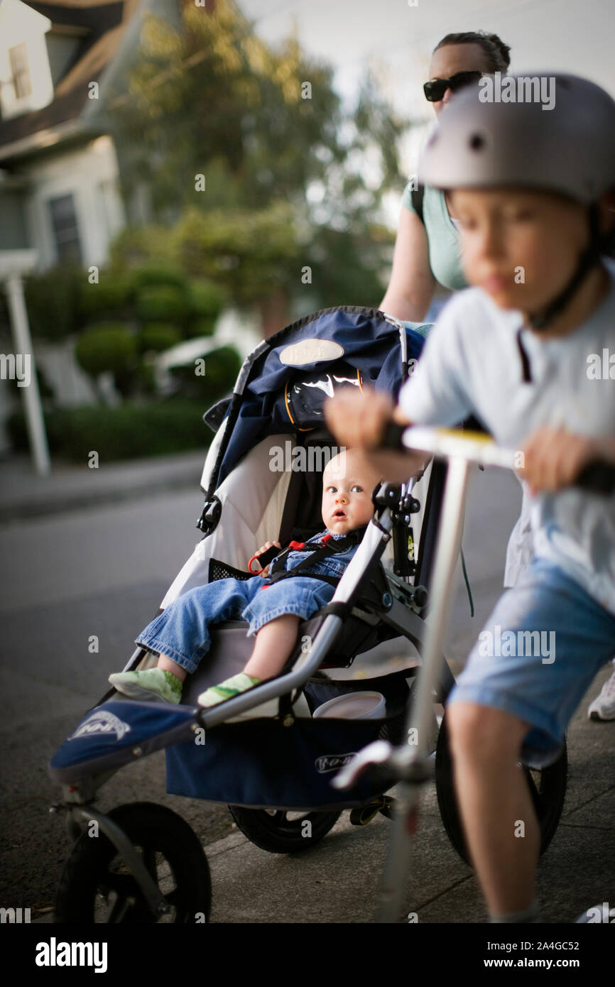 Garçon sur un scooter avec ses mi-mère adultes poussant son tout-petit frère dans un landau à l'extérieur. Banque D'Images