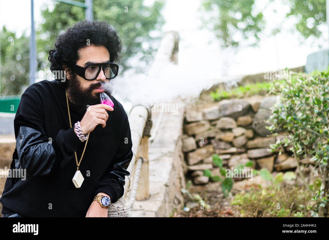 Vape l'homme. Portrait de plein air d'un jeune homme blanc brutal avec une grande barbe et des lunettes en bouffées laissant hors de la vapeur provenant d'une cigarette électronique en face de l'ancienne clôture en bois dans le village. Vaping. Banque D'Images