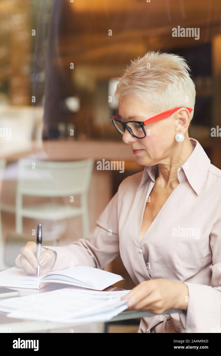 Concentrated mature businesswoman with blonde cheveux courts portant des lunettes assis à la table et d'écrire quelque chose dans les documents in cafe Banque D'Images