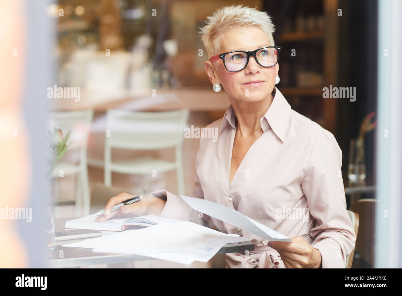 Femme mature blonde avec des cheveux courts en lunettes assis à la table et l'utilisation de documents in cafe Banque D'Images